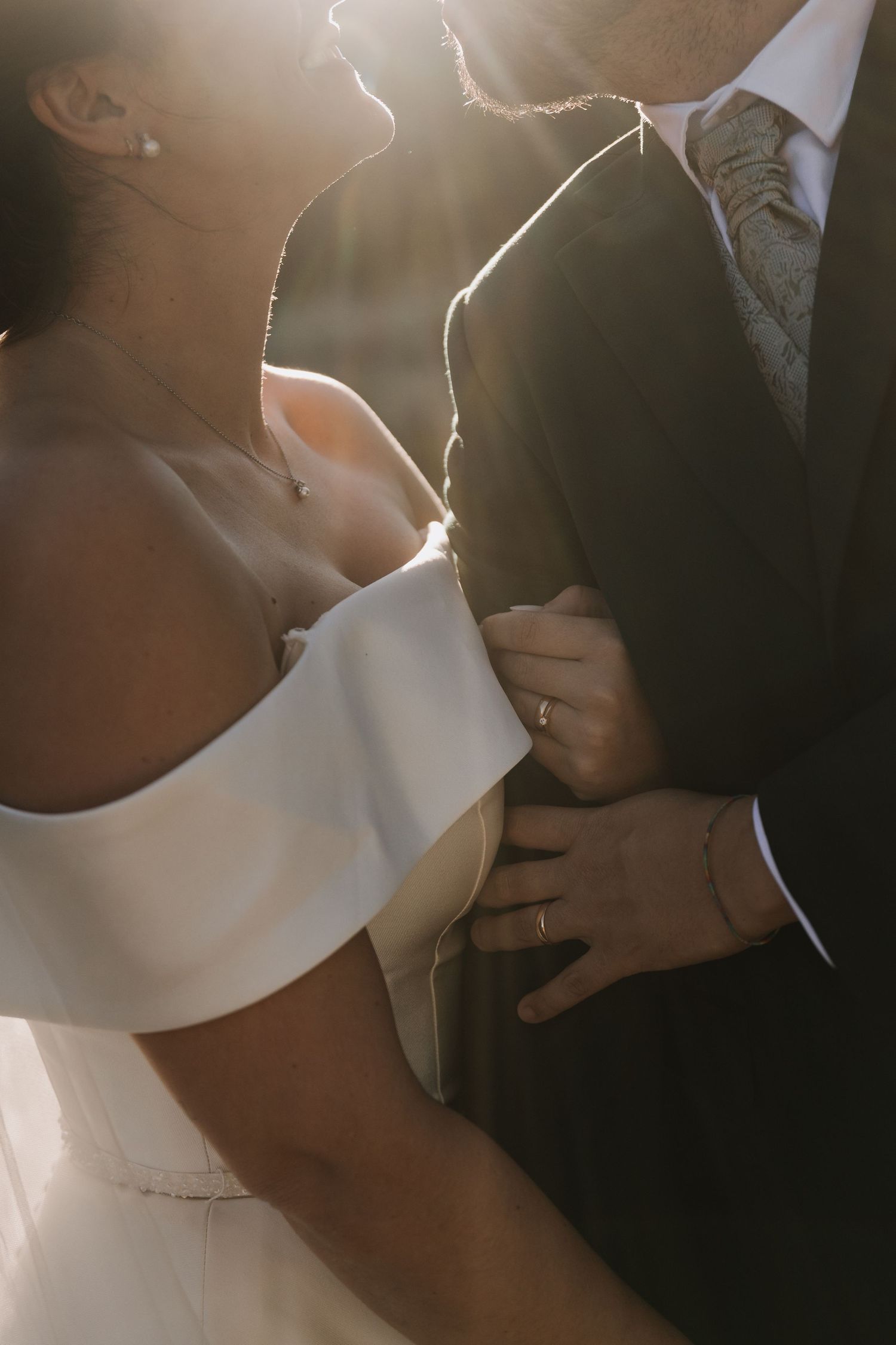 Close-up of bride in off-shoulder gown embracing groom in suit, both bathed in warm golden sunlight. Destination wedding