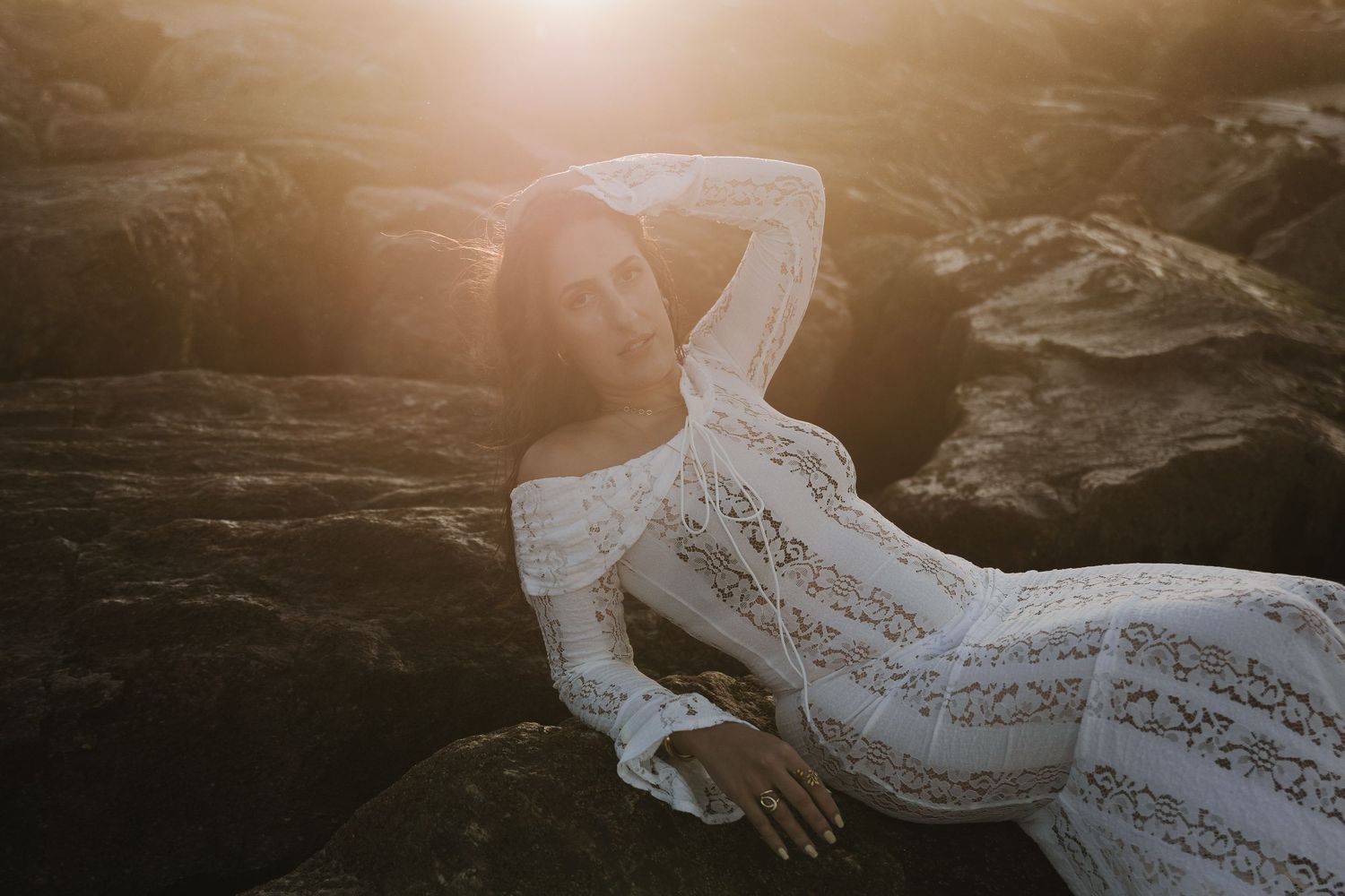 A woman in a white lace dress reclines on rocky terrain bathed in warm golden sunset light.