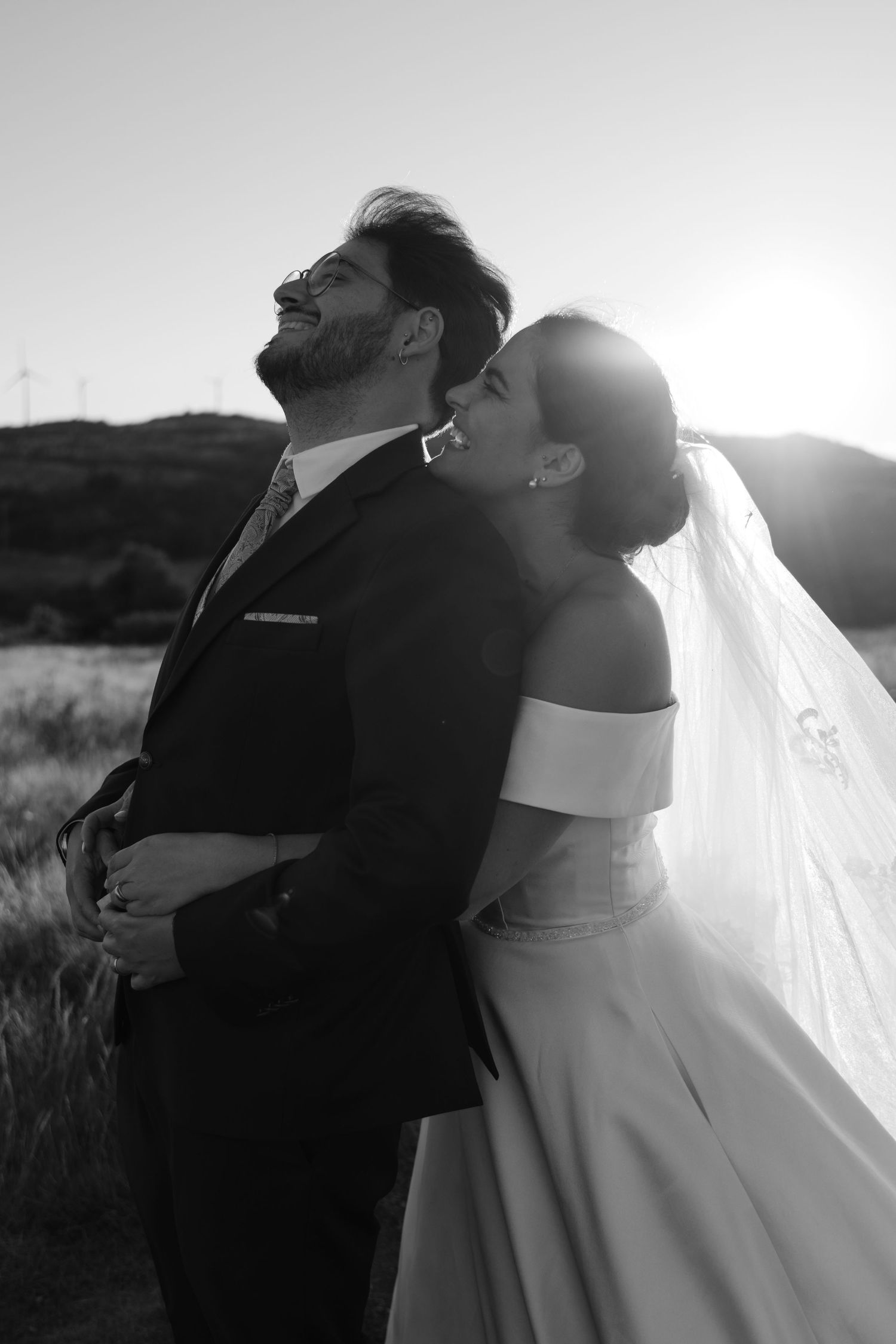 Black and white photo of bride kissing groom on cheek as he laughs joyfully at sunset in a field.