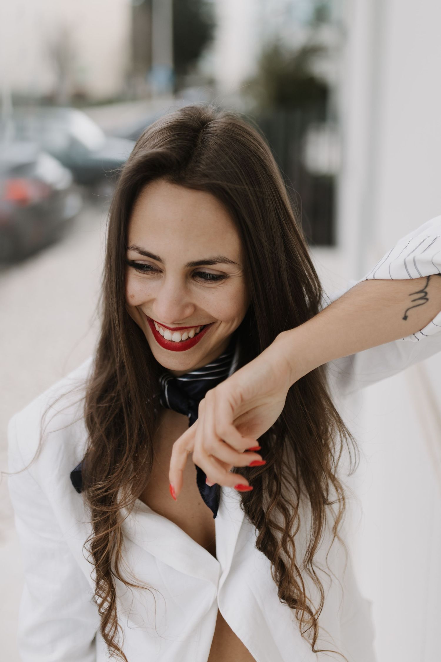 Laughing brunette woman with red lips and tattoo wearing white shirt in urban outdoor setting.