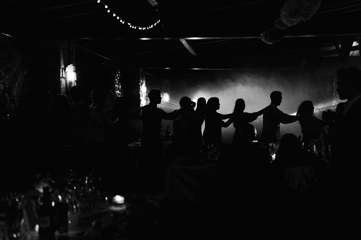 Silhouetted wedding party holding hands on a dimly lit dance floor in a black and white photo.