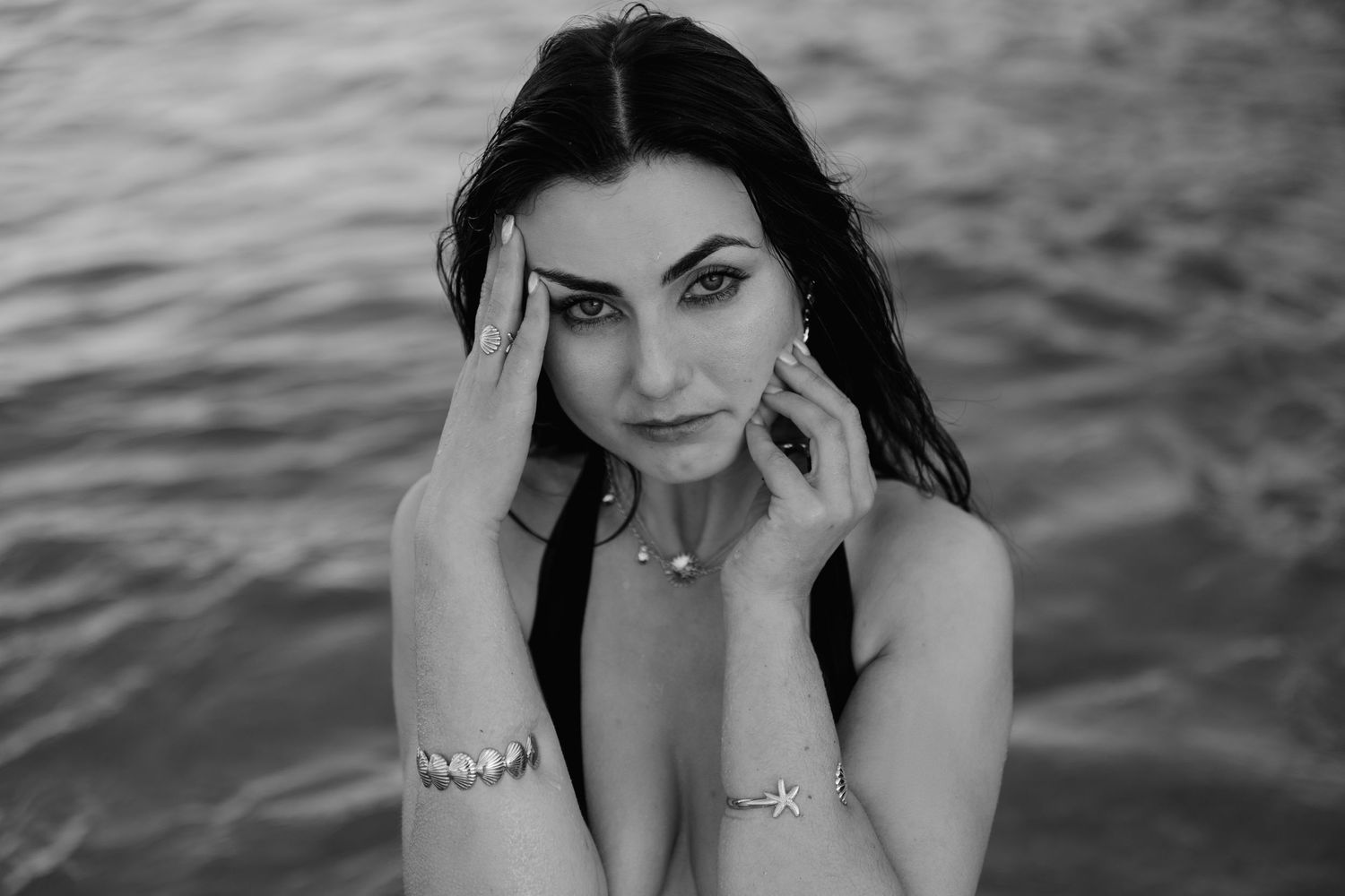 Black and white close-up portrait of a dark-haired woman with striking eyes posing near shimmering water.