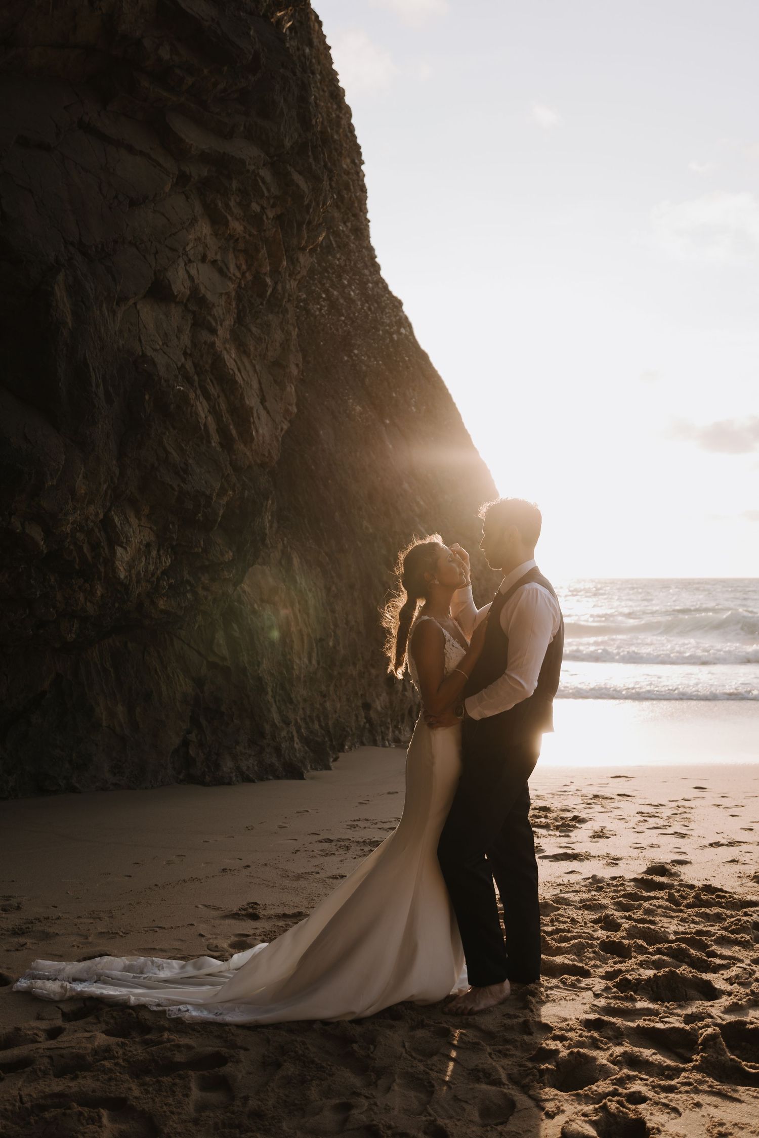 Bride and groom embracing on a sandy beach at golden sunset beside a dramatic rocky cliff.