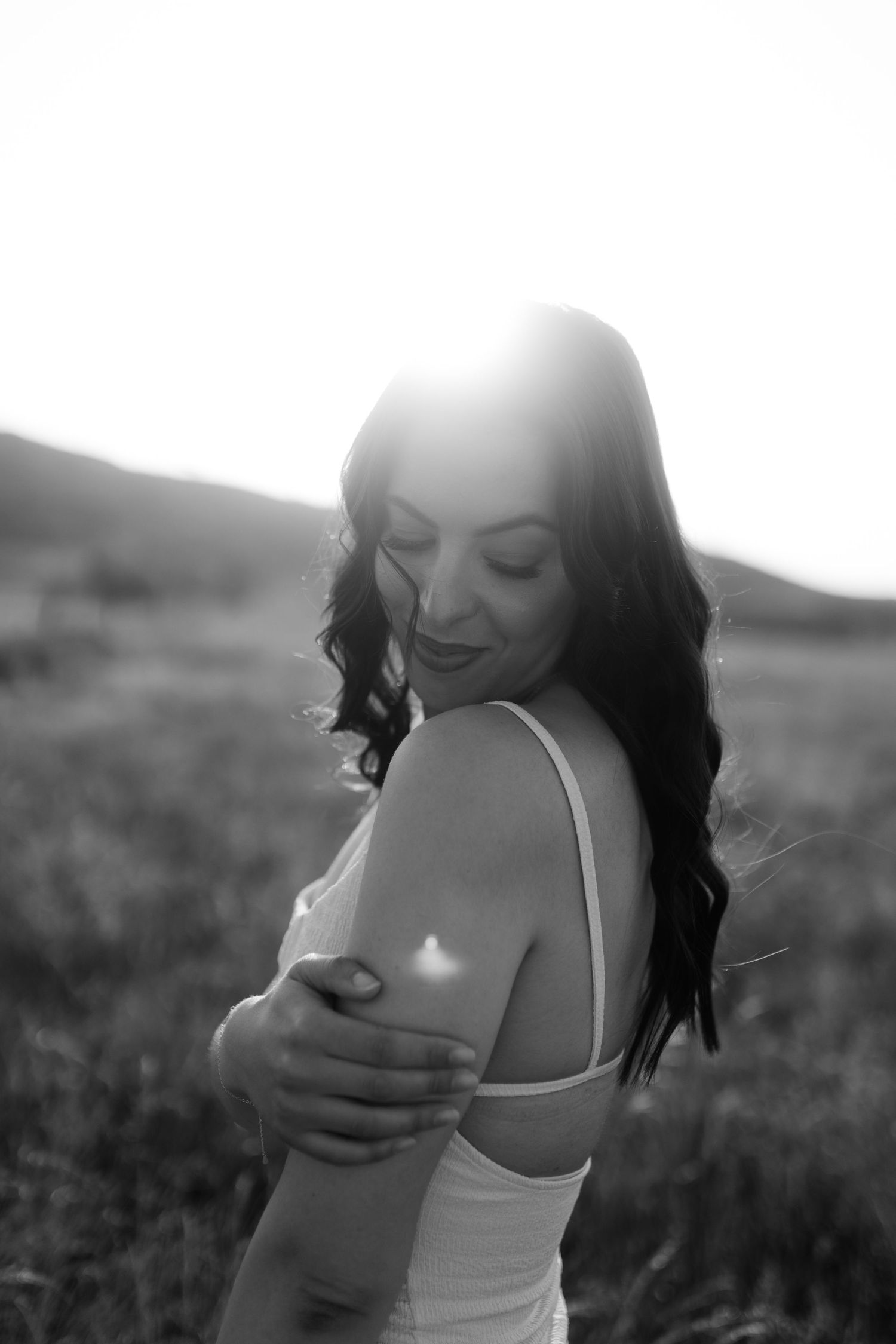 Black and white portrait of smiling brunette woman in white dress with sunlit field background.