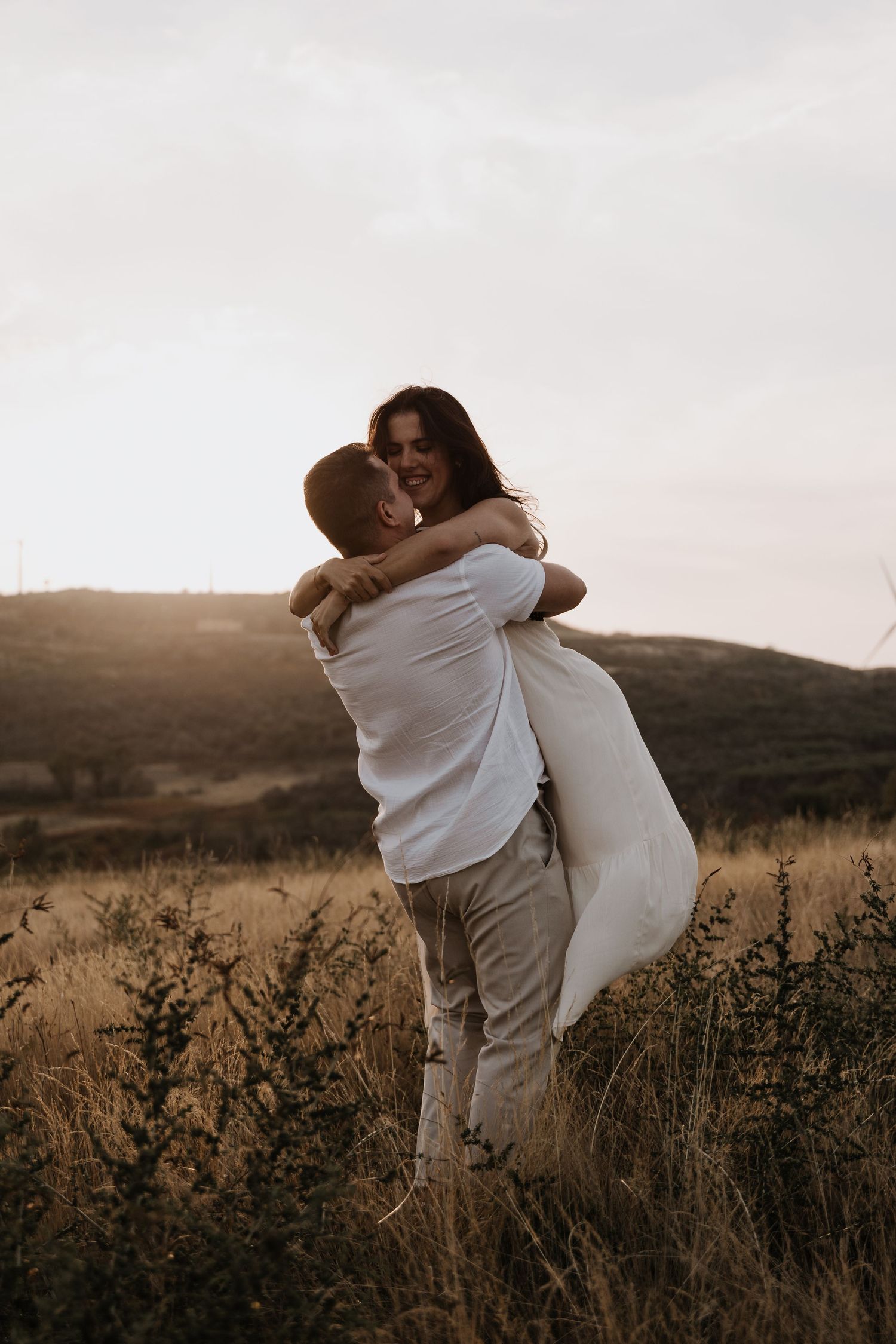 Couple Photoshoot with a man lifts and embraces a laughing woman in a white dress in a golden sunset field with rolling hills.