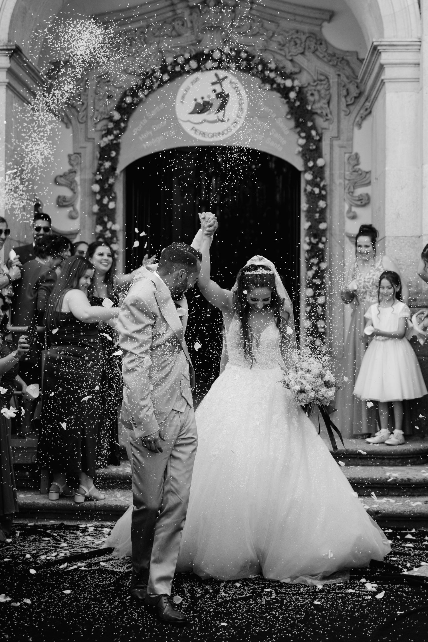 Black and white photo of newlyweds exiting church as guests shower them with confetti in celebration.