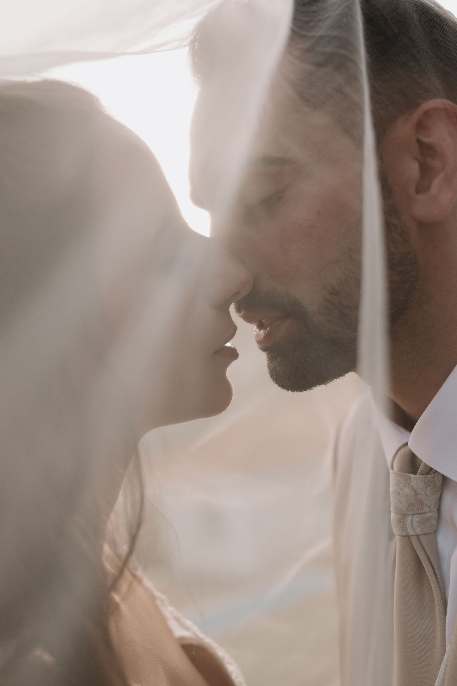 Intimate close-up of bride and groom nearly kissing under a flowing veil in soft, ethereal light.