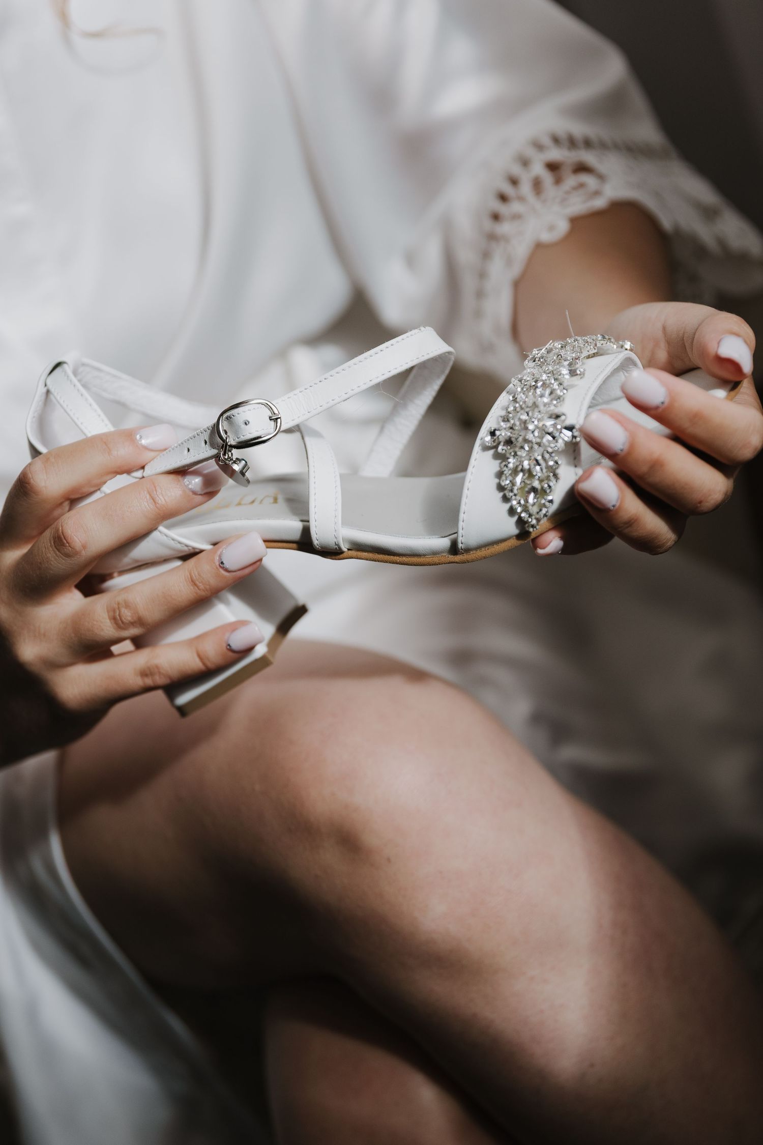 Bride holding embellished white wedding sandal with crystal brooch detail while getting ready.