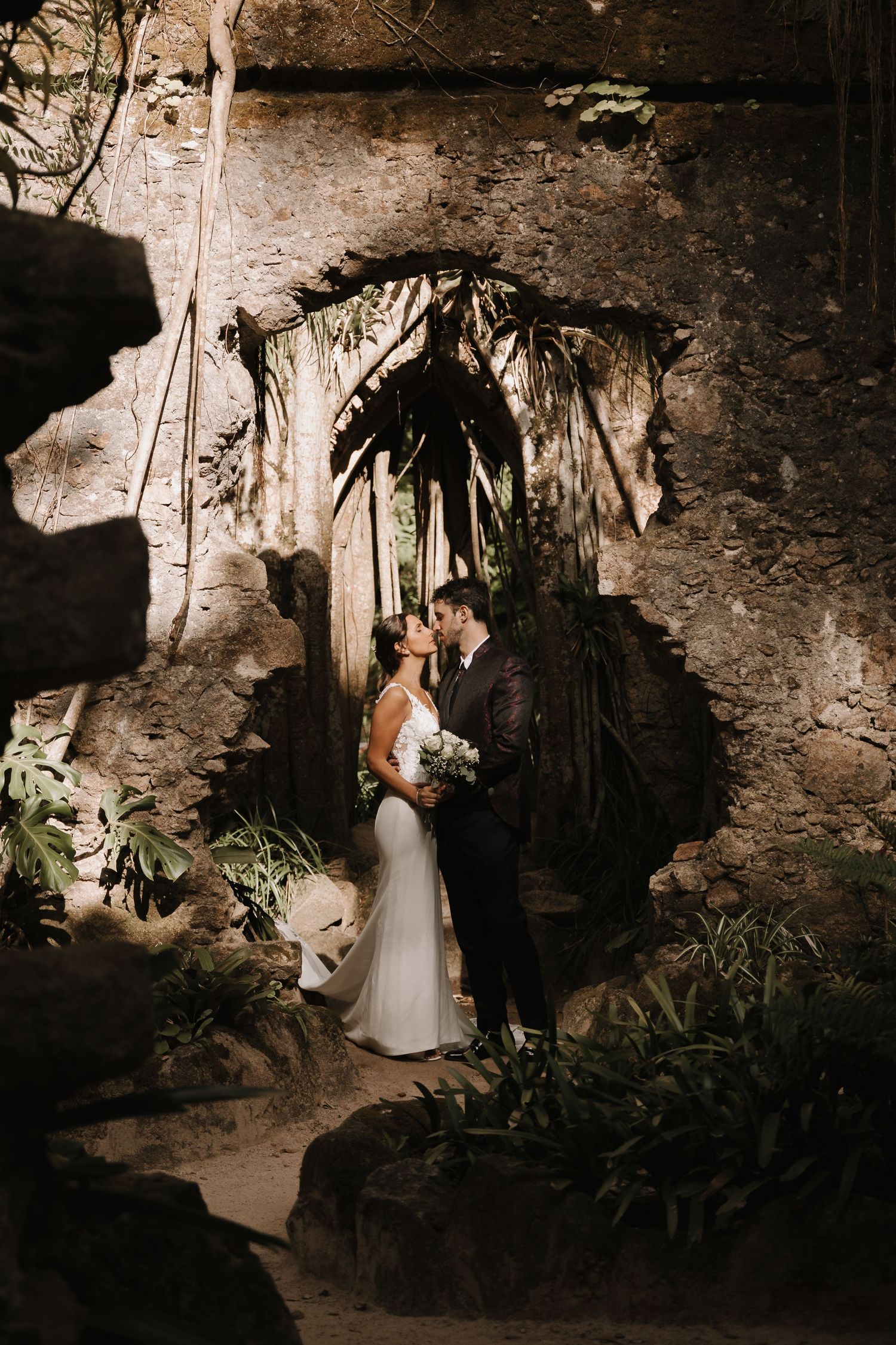 Bride and groom sharing a kiss in a dramatic cave-like setting surrounded by tropical foliage.