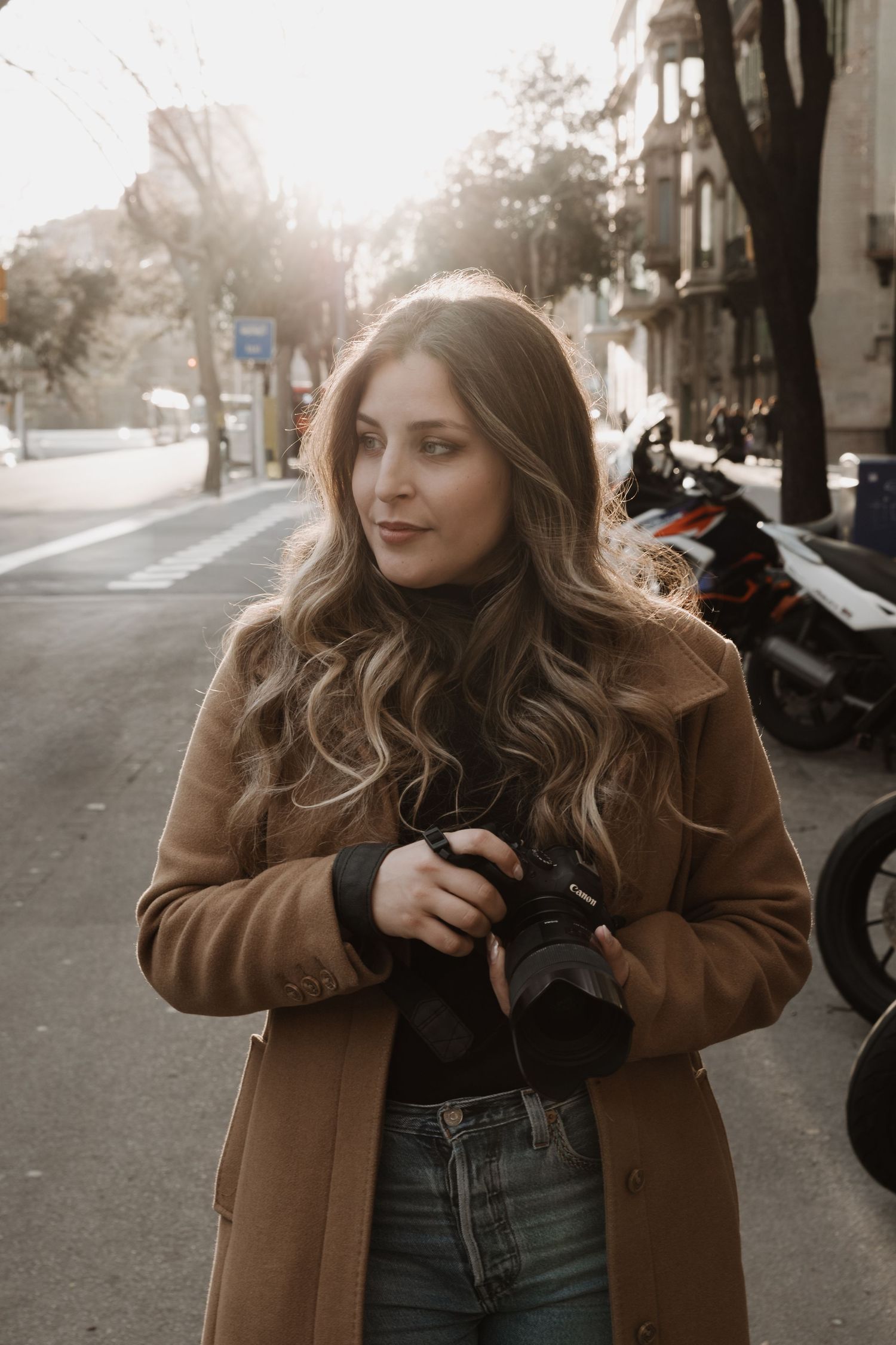 Young woman with wavy hair holding a camera on a sunlit city street wearing a camel coat.