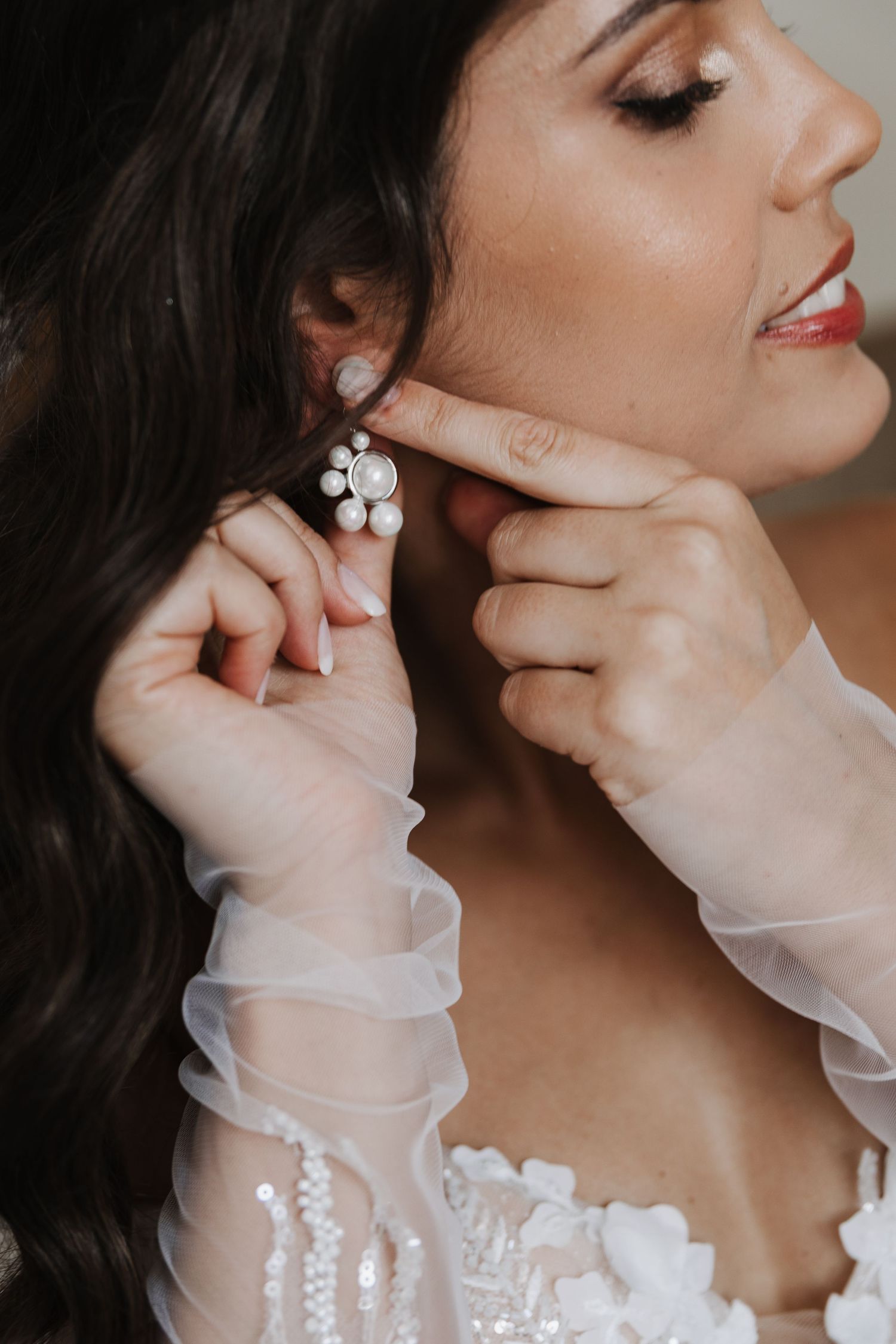 Close-up of bride with dark hair putting on elegant pearl earrings in wedding dress.