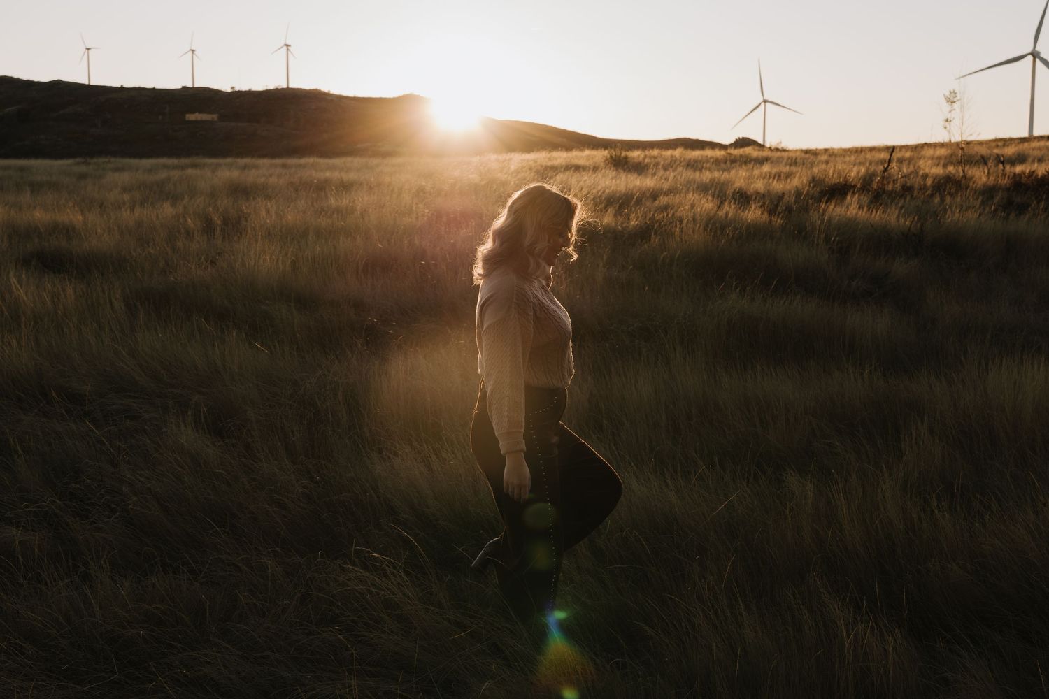 Woman walking through golden grass field at sunset with wind turbines glowing in background.