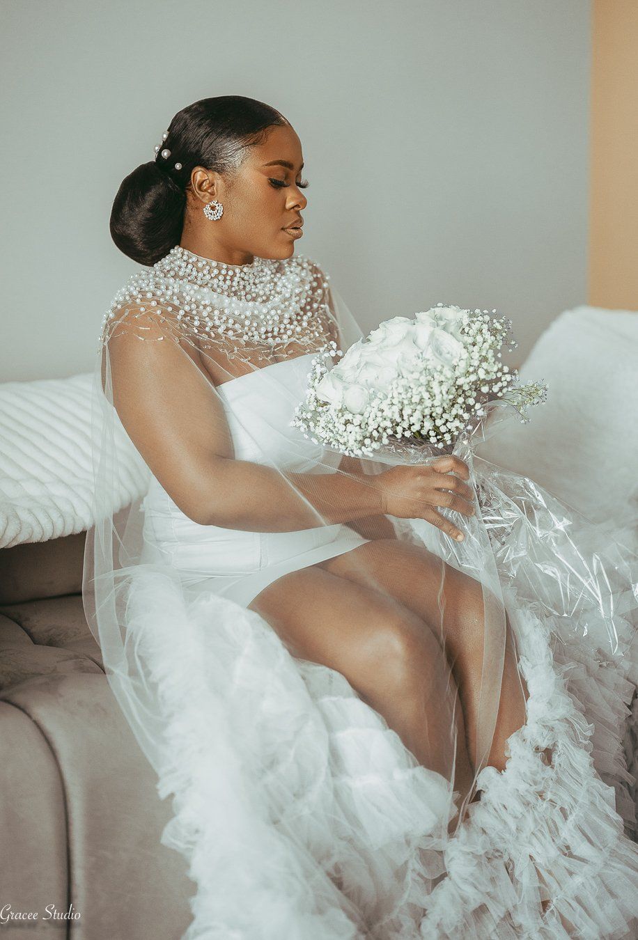 A bride in a luxurious white gown with crystal embellishments reclines elegantly on a beige couch during a photoshoot.