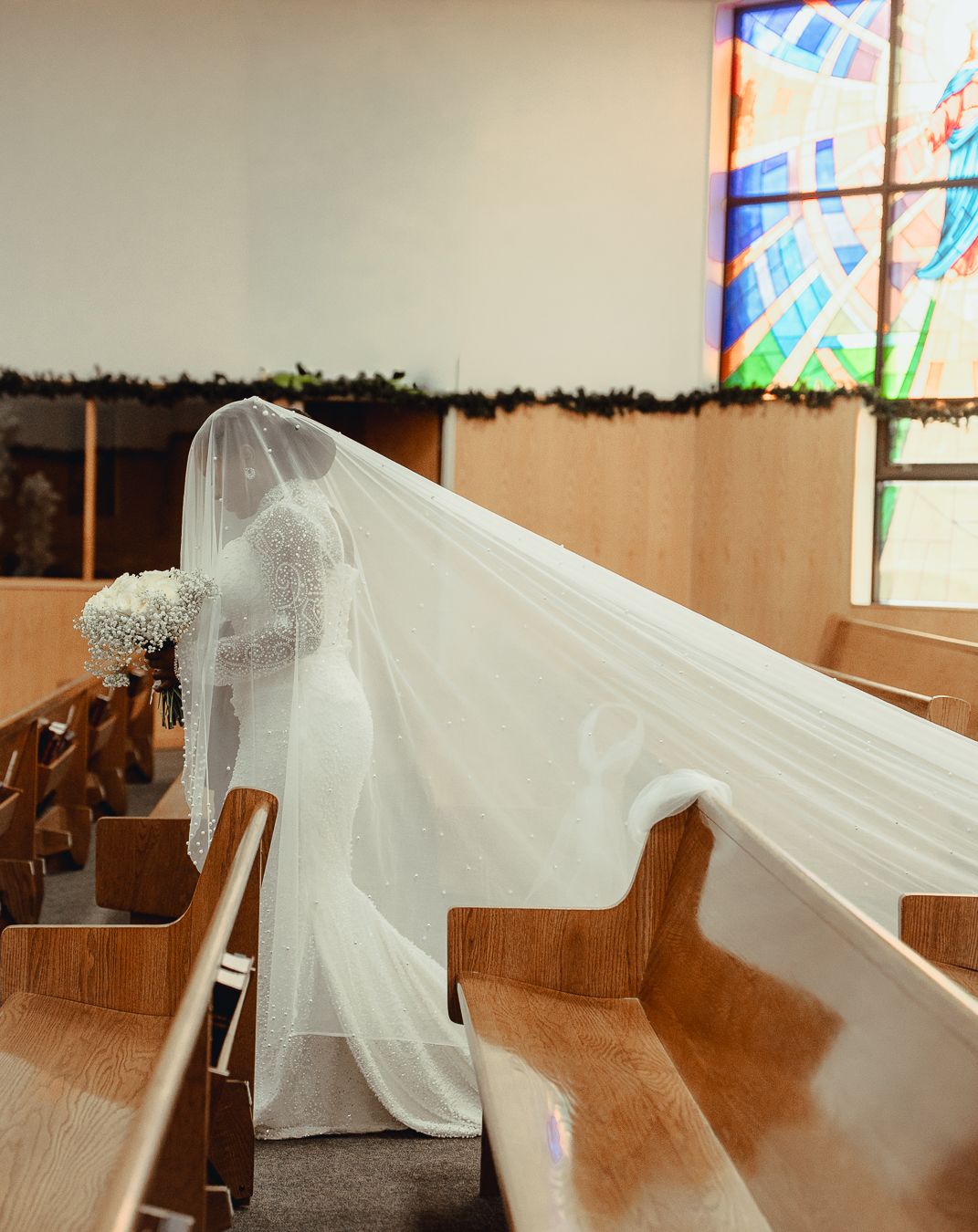 A bride wearing flowing white wedding veil drapes elegantly across wooden church pews with stained glass windows visible in the background.