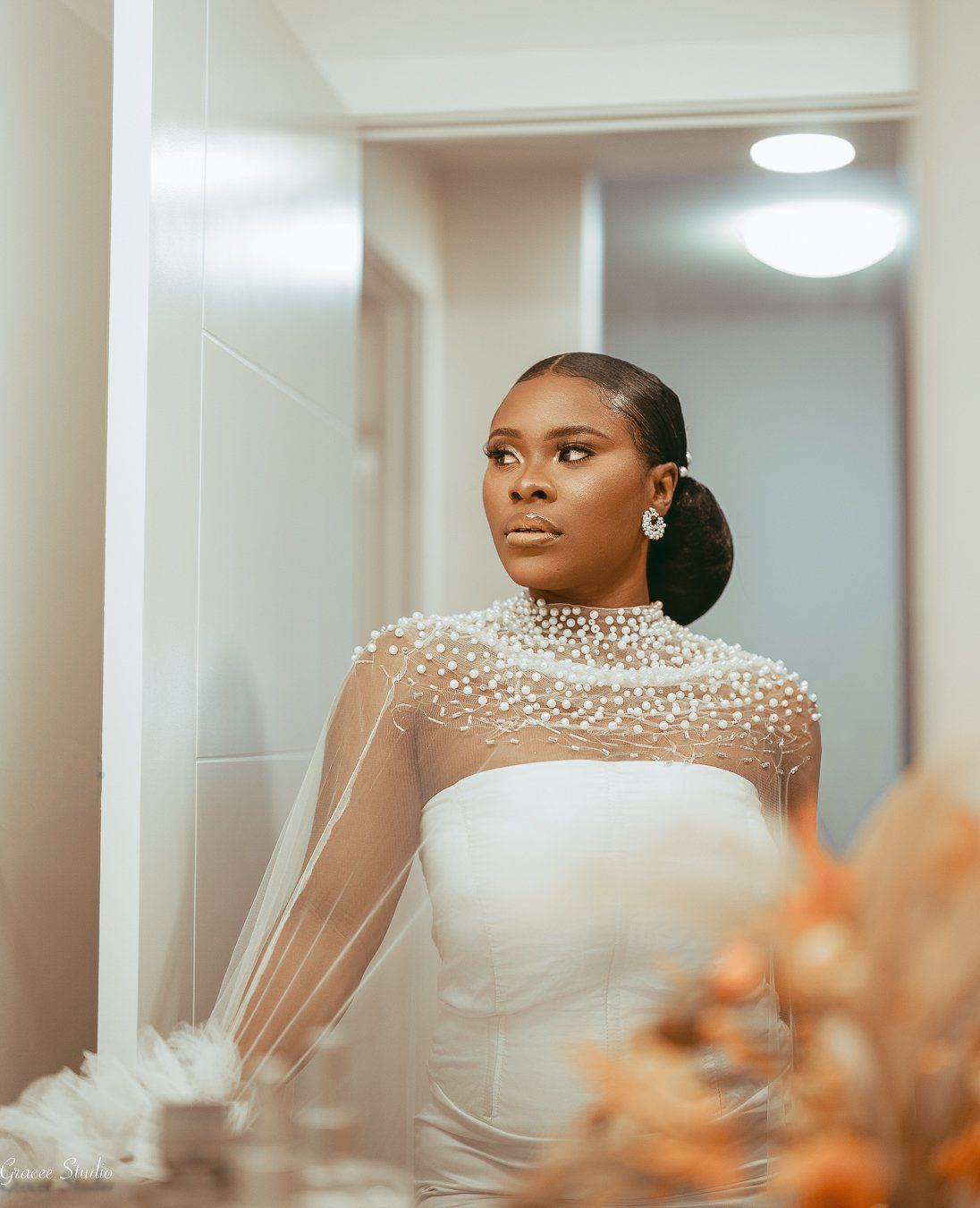 Bride in elegant white gown with pearl-embellished neckline and sheer sleeves poses in bridal suite.