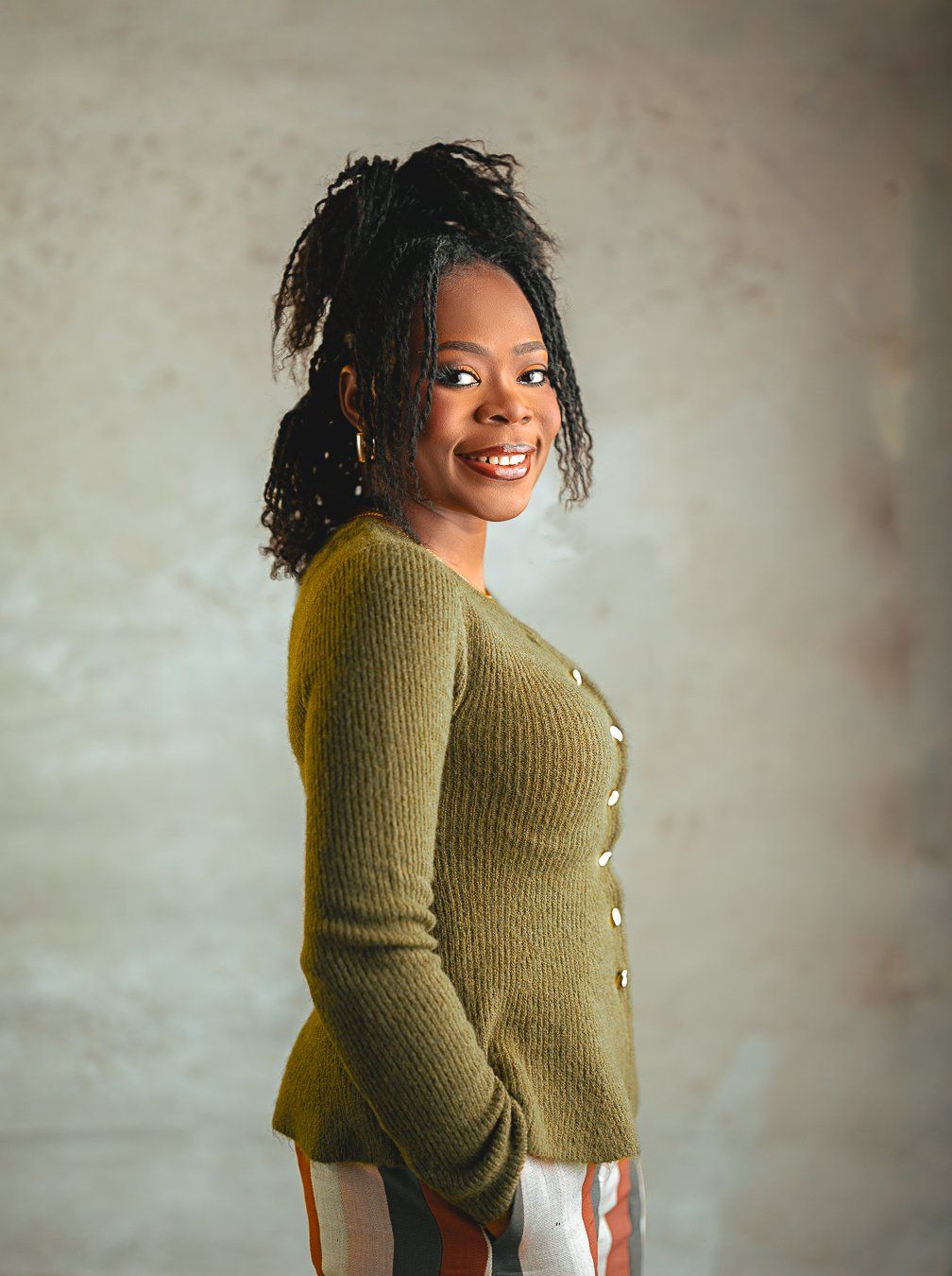 A lady in an olive green cardigan sweater poses against a neutral wall with a joyful expression.
