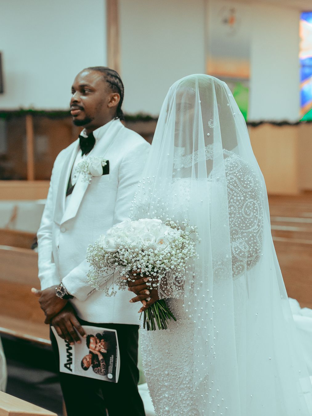 A bride holding a delicate baby's breath bouquet stands beside her groom in a white suit during a church wedding ceremony.