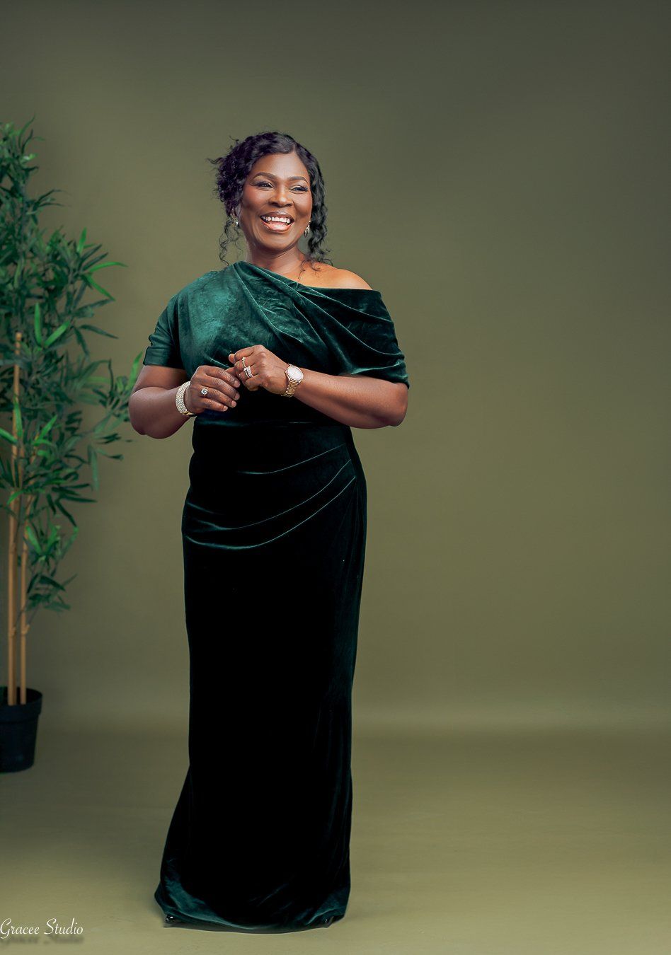A woman in a green velvet dress poses elegantly against a sage green backdrop with bamboo plants in the studio setting.