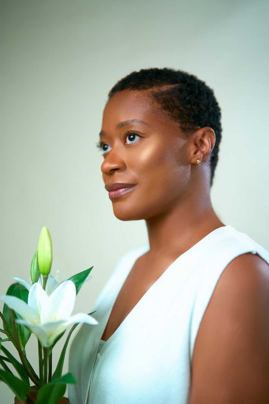 Side profile portrait of a lady wearing white sleeveless top holding white lily flowers against soft green background.