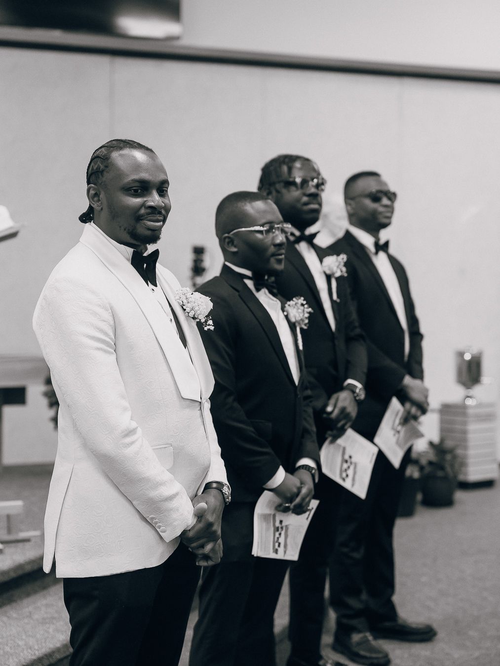 A group of groomsmen in formal attire stand together at a wedding ceremony, with some wearing white and black suits.