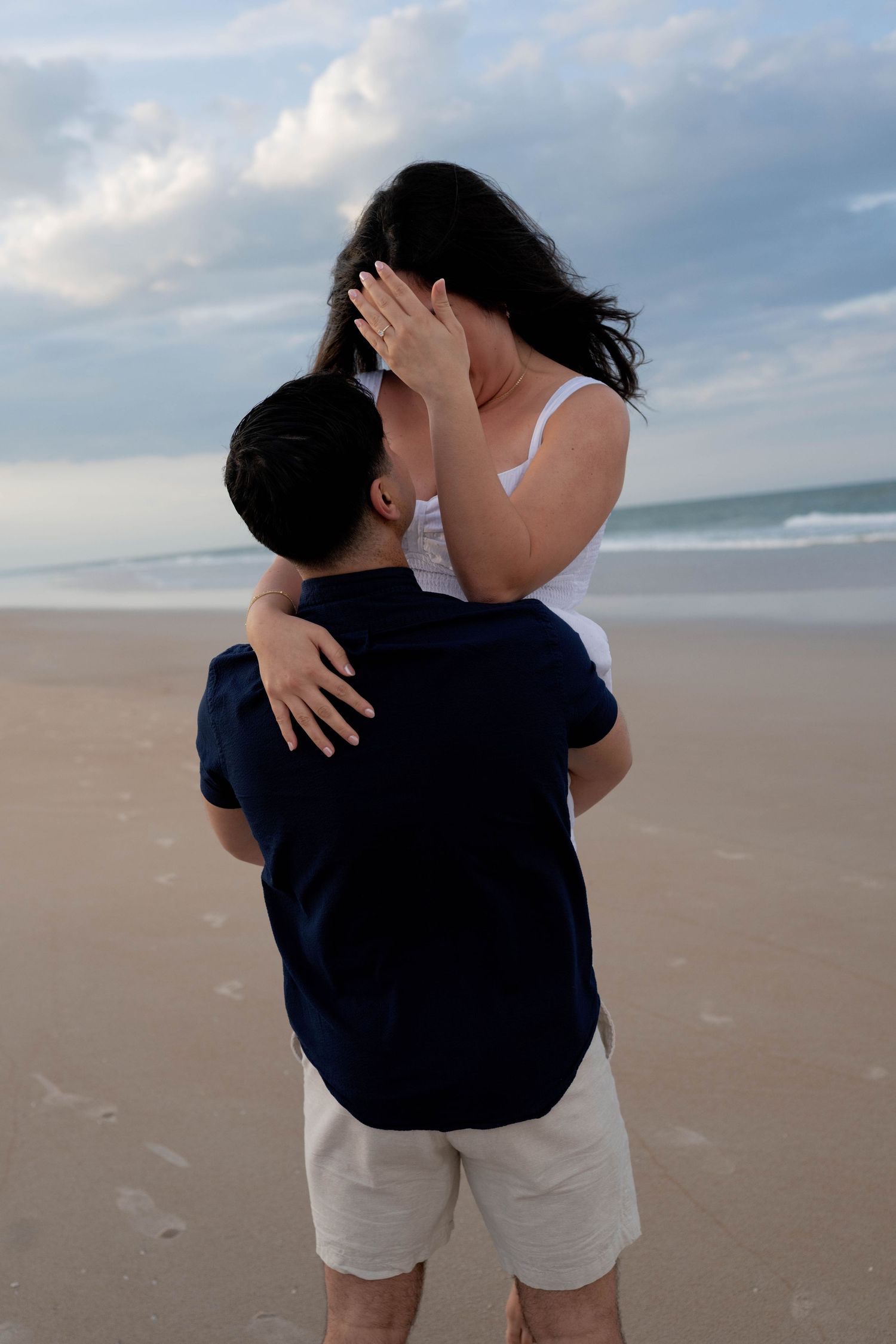 Couple embracing on a sandy beach, woman lifted by man in navy shirt, ocean waves and cloudy sky in background.