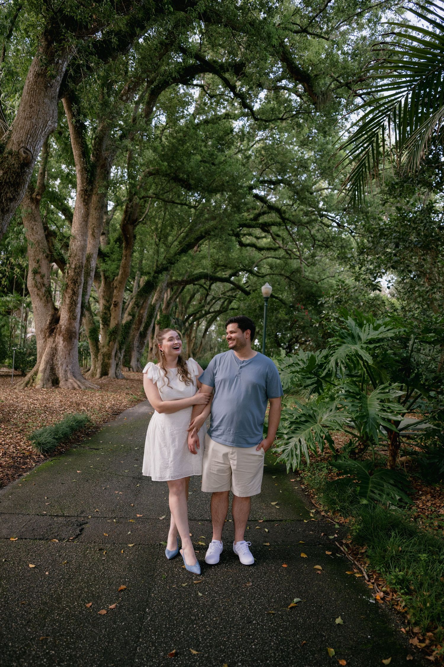 A couple walks hand-in-hand on a tree-lined path, laughing together in a lush green park setting.