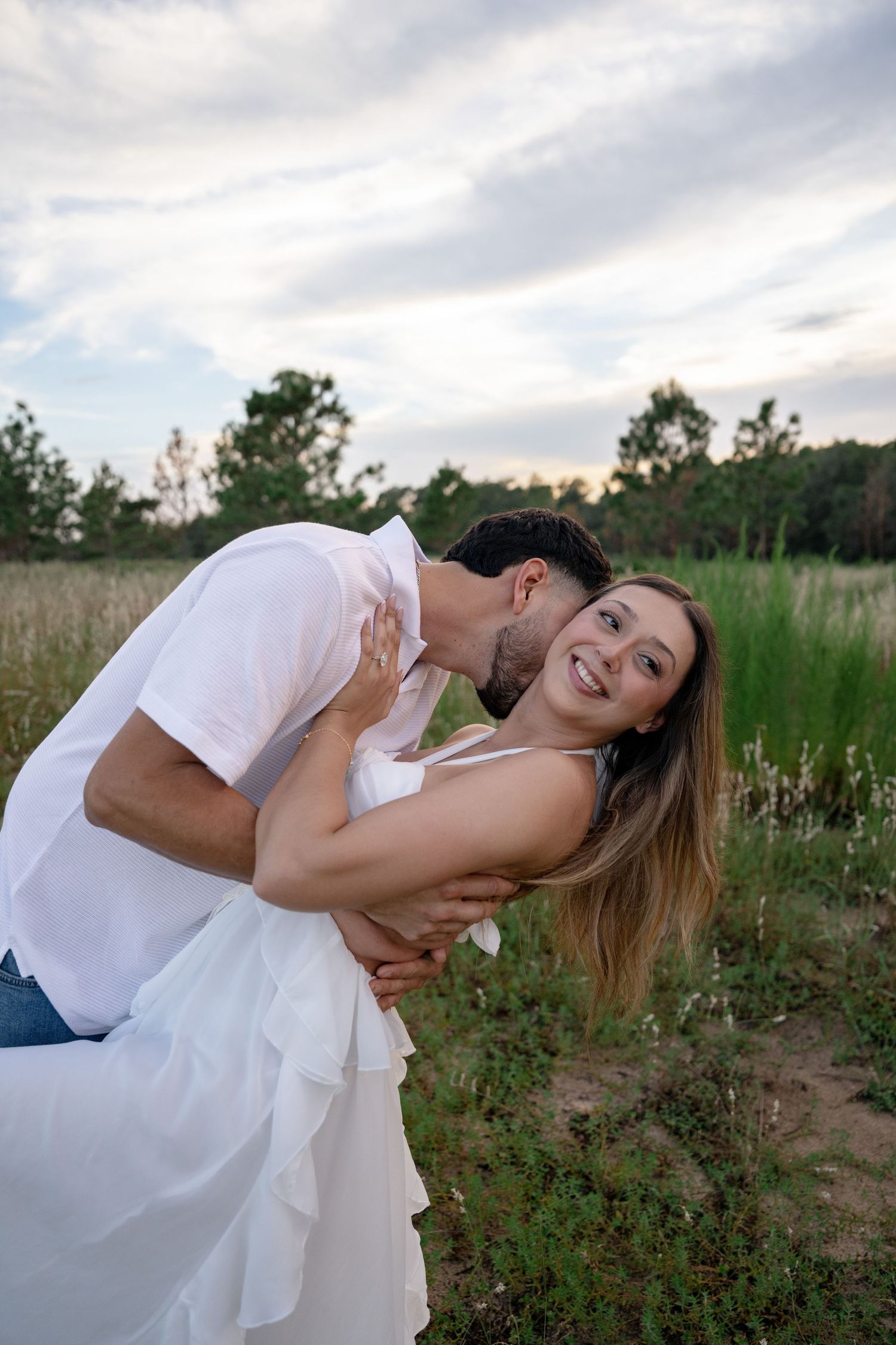 Couple embracing outdoors, man kissing smiling woman in white dress in a green field at dusk.
