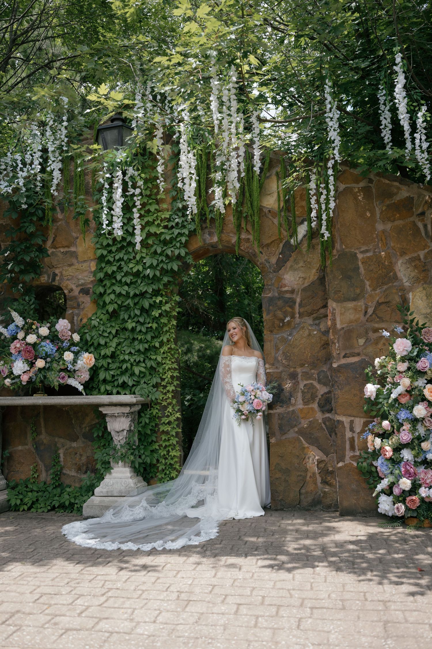 Bride in white gown and veil stands before stone wall with ivy, white wisteria, and colorful floral arrangements.