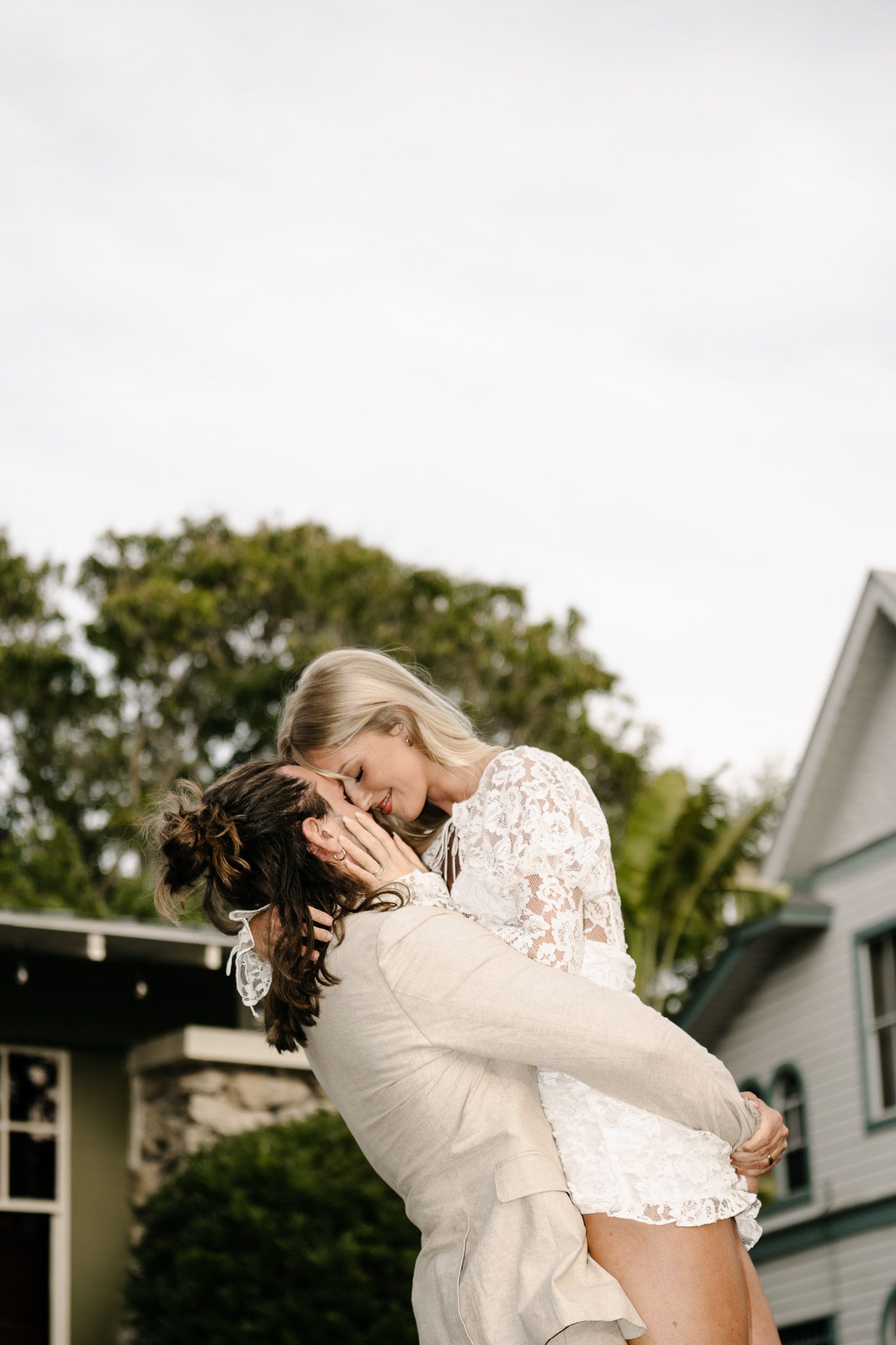 Playful wedding couple embracing on lawn outside white house.