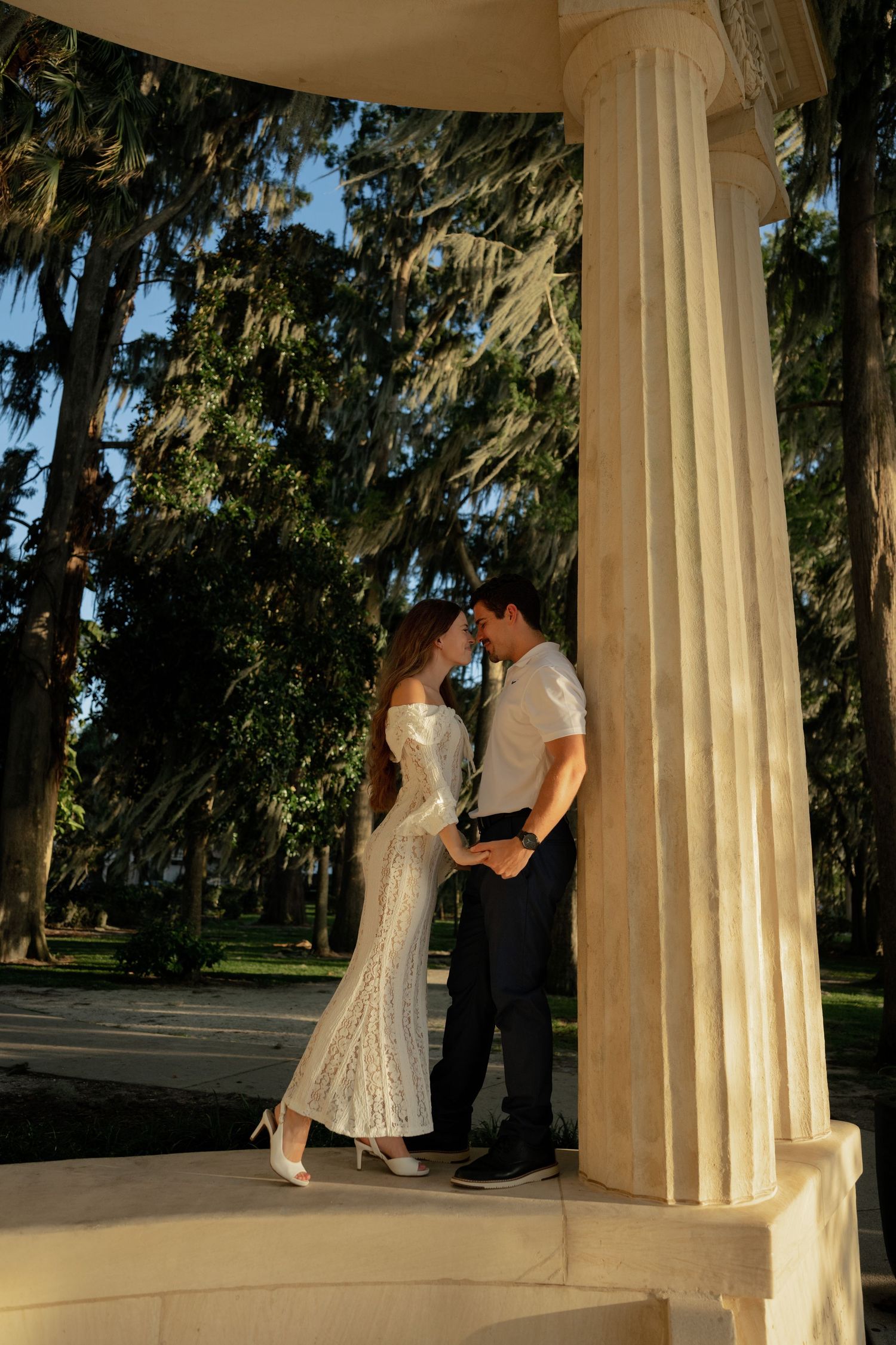 Couple in wedding attire sharing a romantic moment between white columns in a sunlit park with tall trees.