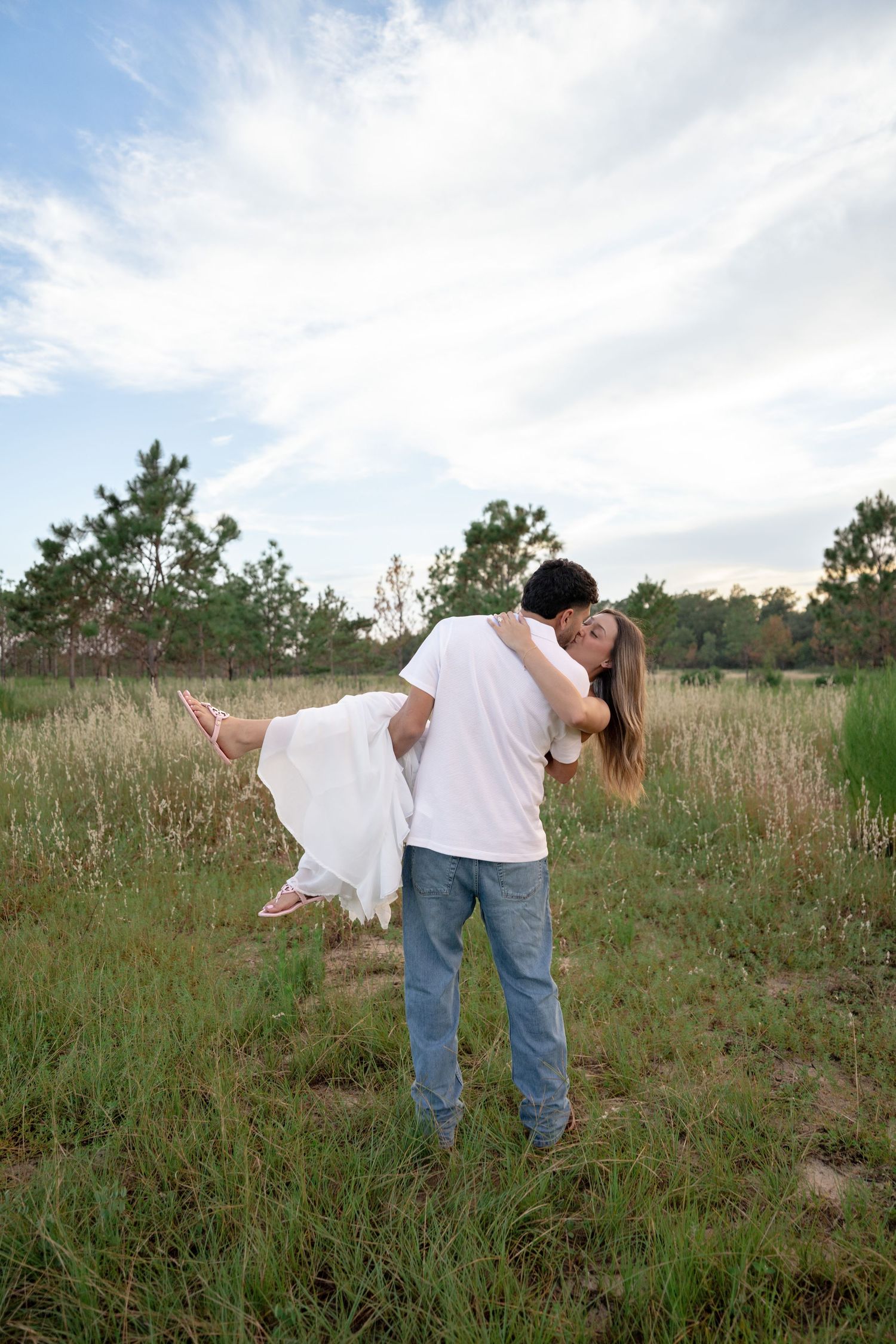 Man in white shirt and jeans lifting woman in white dress while kissing in a green meadow field.