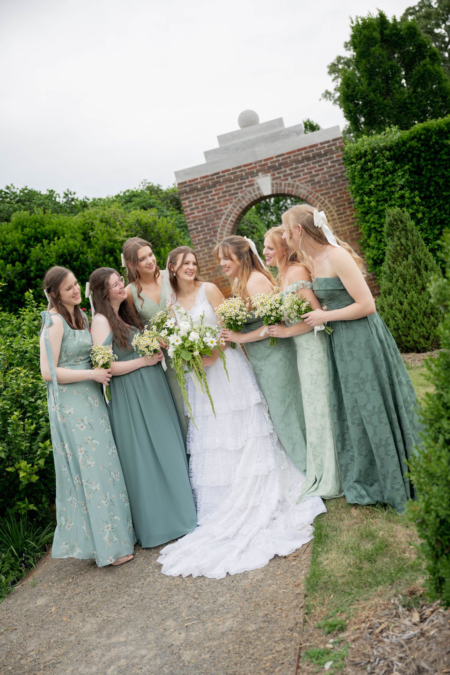 Bride in white gown with six bridesmaids in sage green dresses holding bouquets in a garden setting.