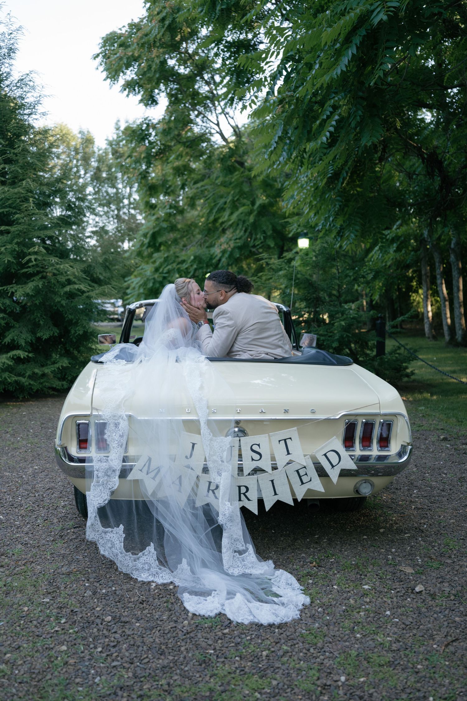 Newlywed couple kissing beside a vintage cream convertible car decorated with a Just Married banner and bridal veil.