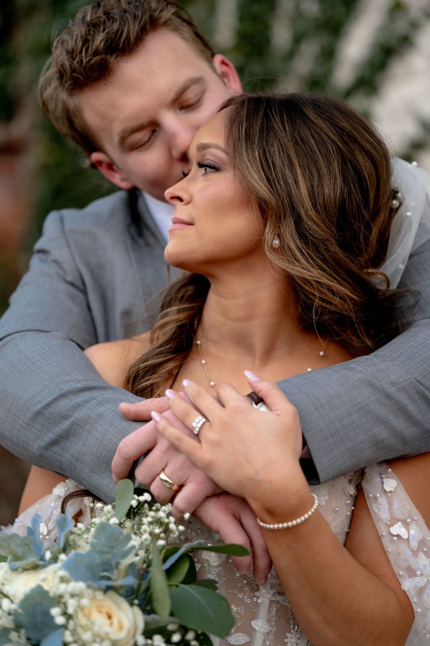 Groom in gray suit embracing bride in lace dress, kissing her cheek while she holds a white and sage bouquet.