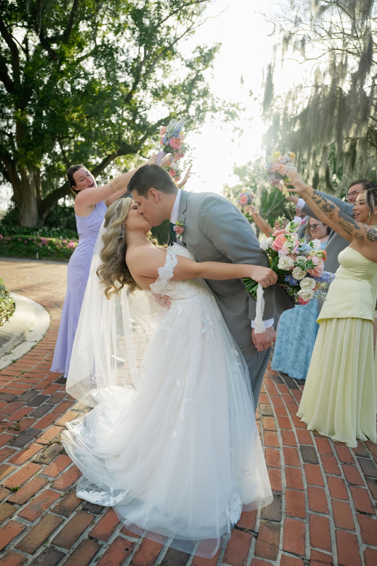 Wedding couple performs romantic dance dip on brick patio surrounded by guests in pastel dresses.