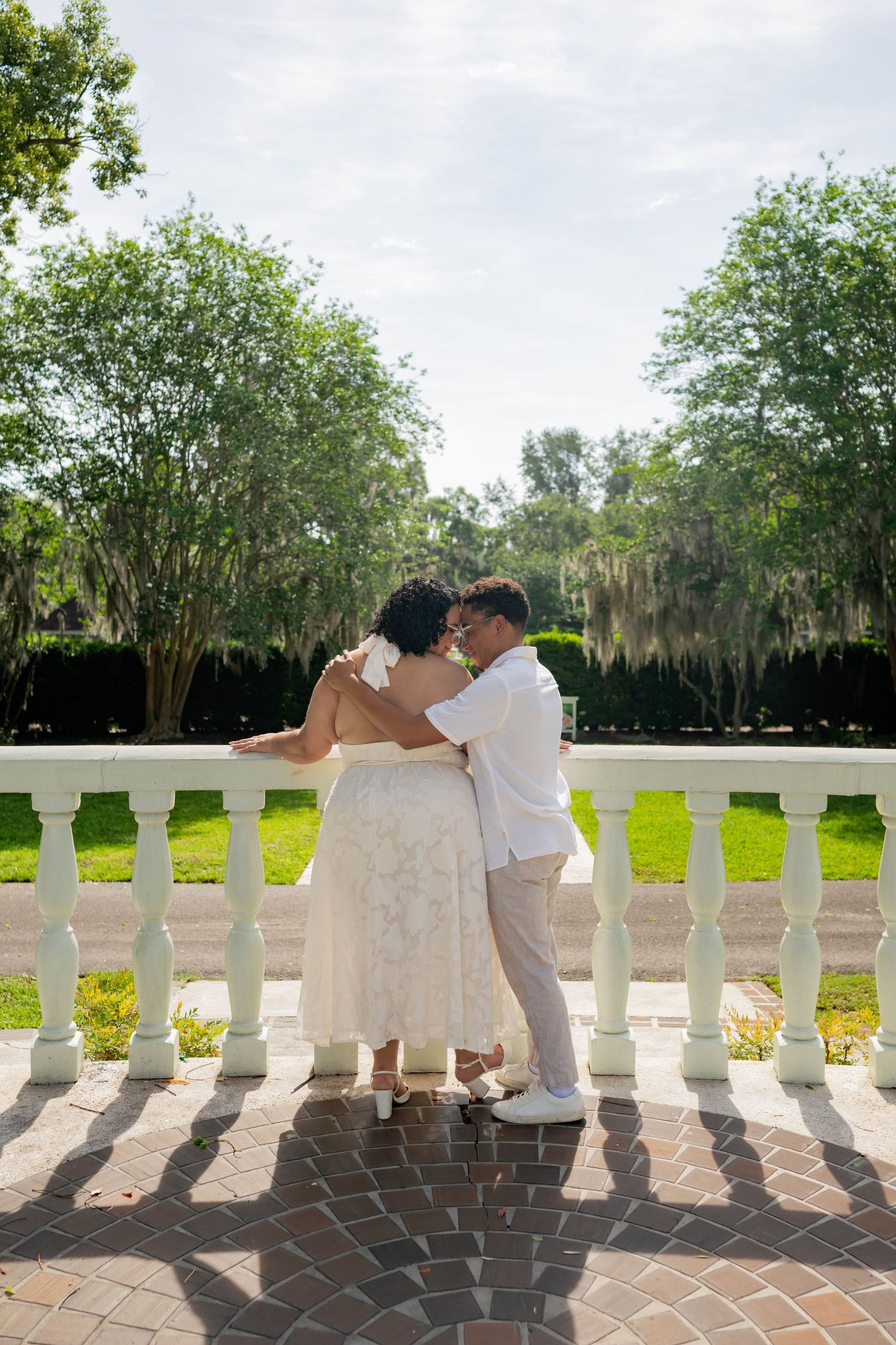 Couple sharing a romantic kiss on a white balustrade terrace surrounded by lush green trees on their wedding day.
