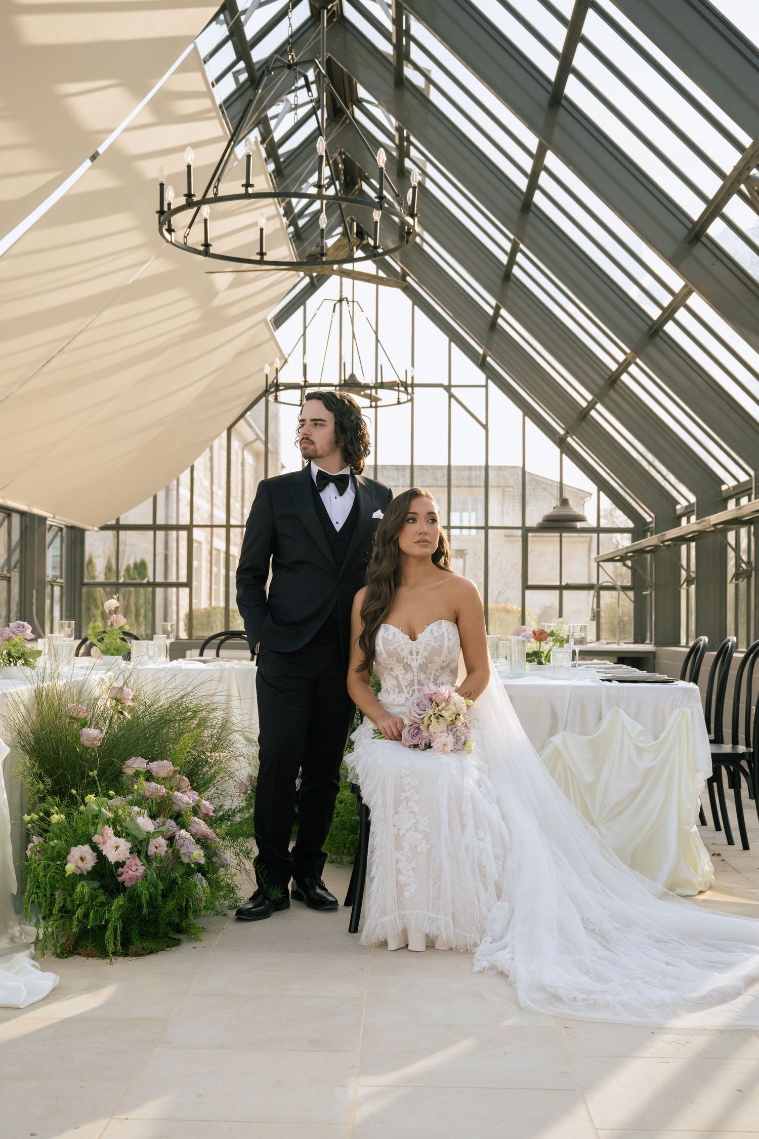 Bride and groom pose elegantly in a sunlit greenhouse wedding venue with floral arrangements and draped ceiling.