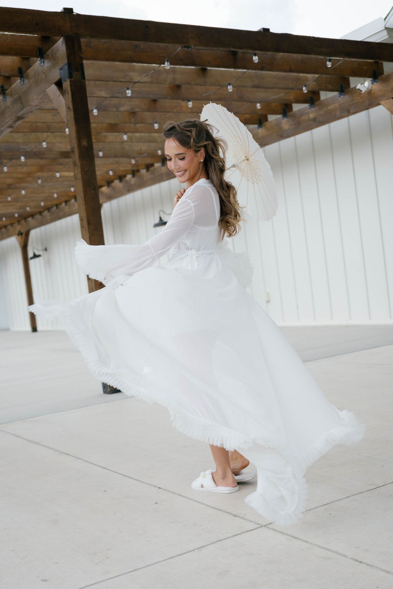 Bride in flowing white gown twirling with a lace parasol outdoors near a rustic wooden pergola.