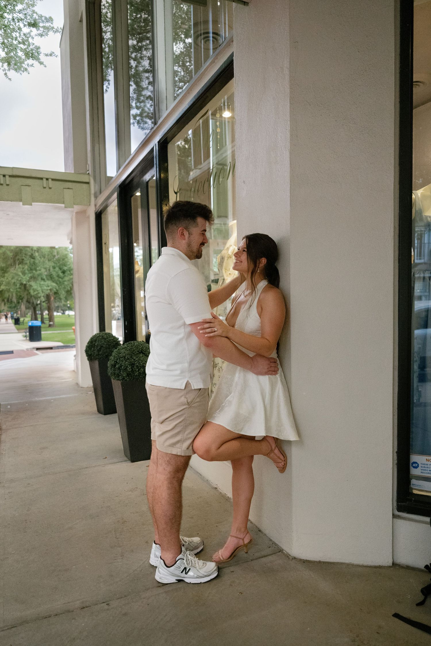 Couple in white outfits share a romantic moment leaning against a building wall on a sunny sidewalk.