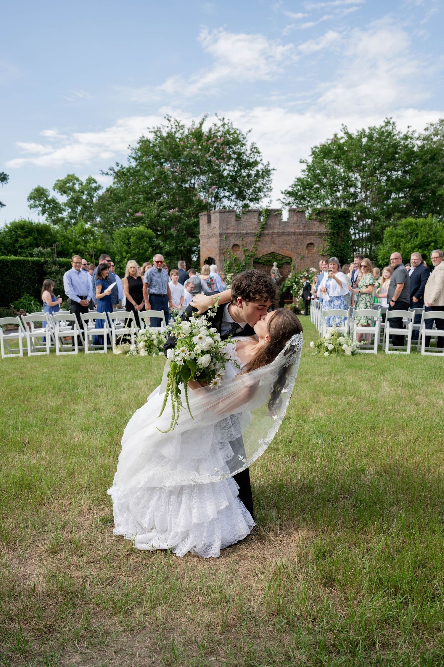 Bride and groom share a romantic dip kiss at outdoor wedding ceremony with guests and brick ruins in background.