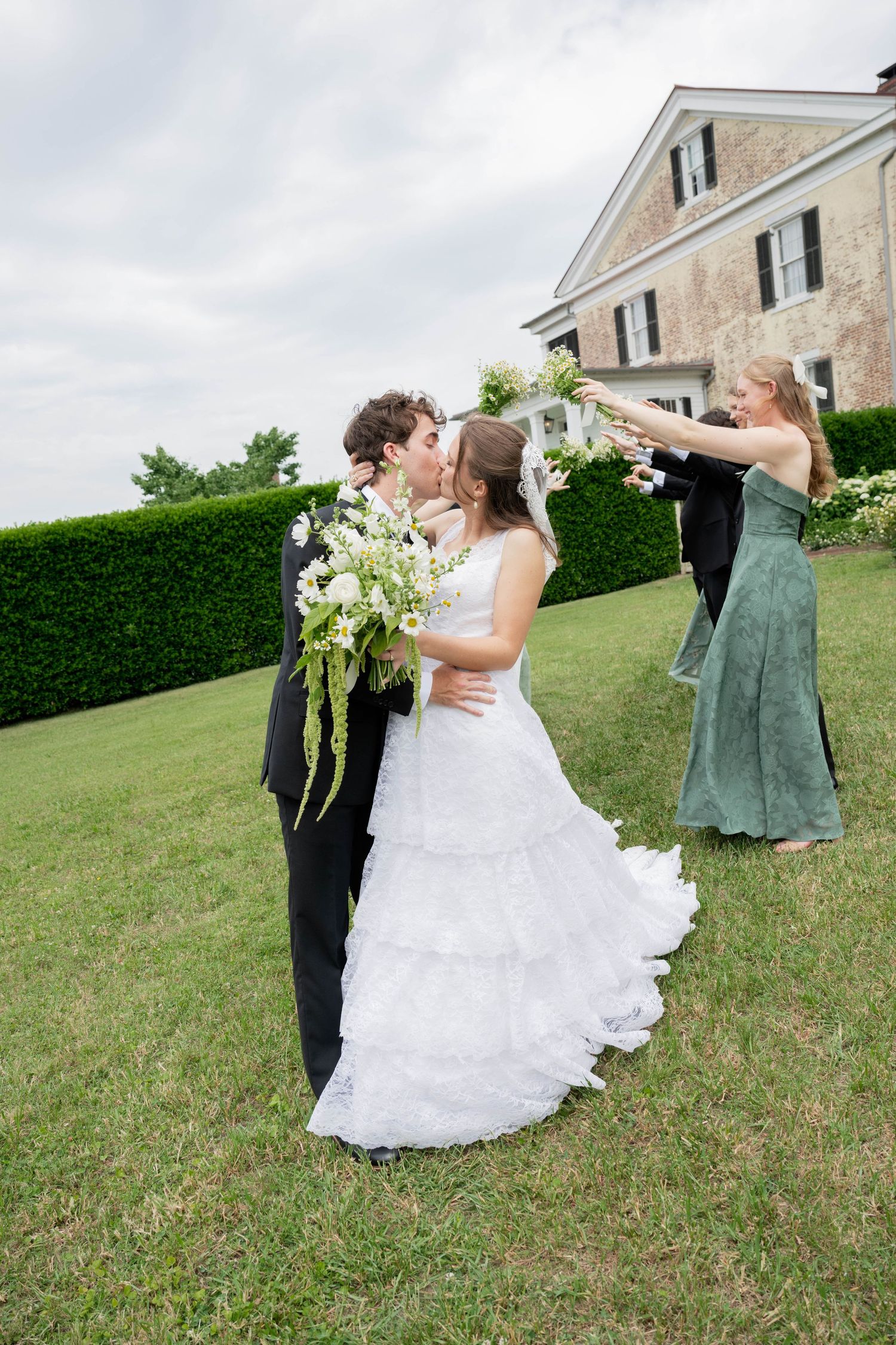 Bride and groom share a kiss outdoors while bridesmaid raises bouquet in celebration at elegant estate wedding.