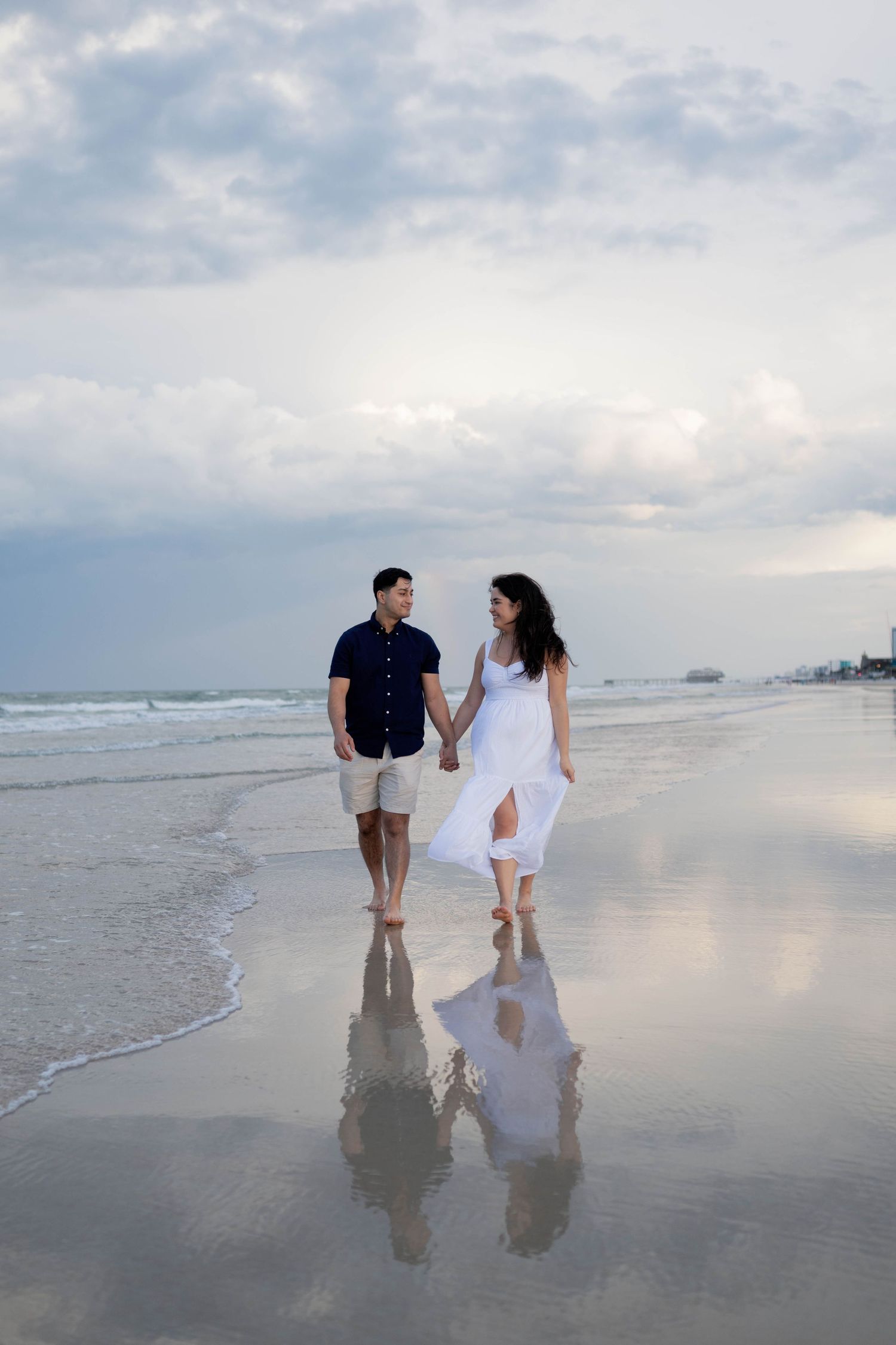 Couple walking hand-in-hand along a reflective wet beach shoreline at sunset, woman in flowing white dress.