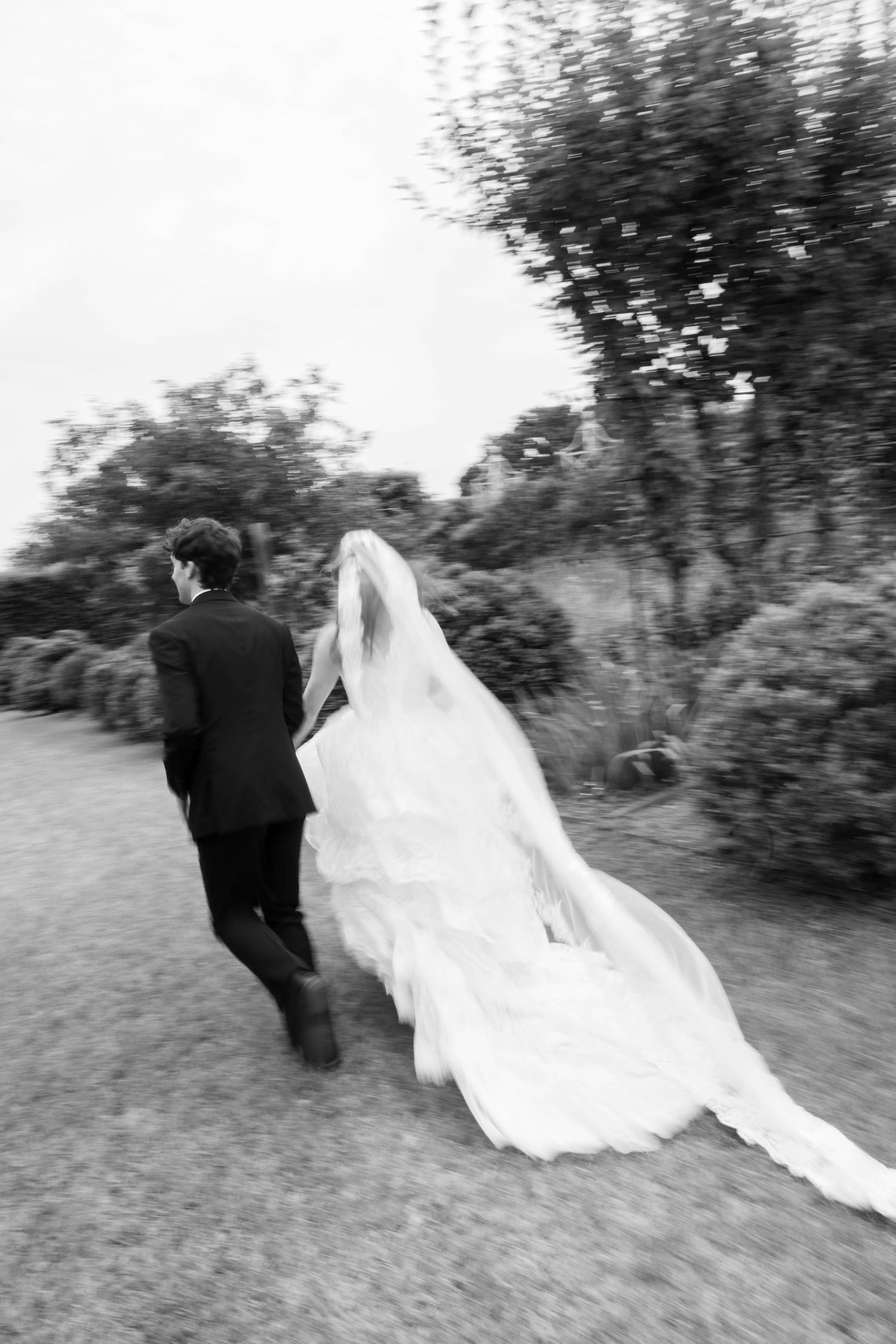 Black and white photo of bride and groom walking away through garden, wedding dress trailing behind them.