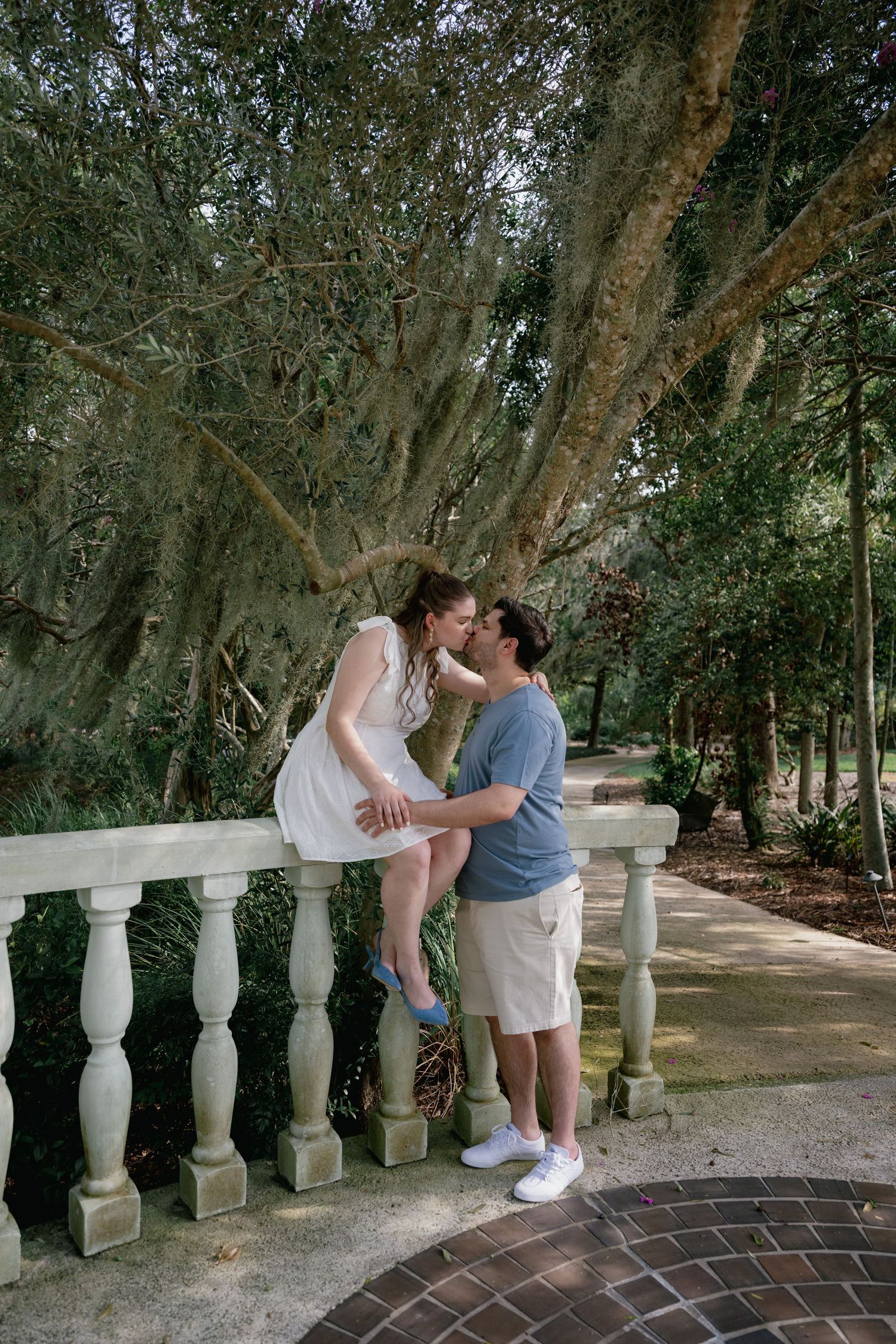 Couple sharing a romantic kiss on a decorative balustrade bridge surrounded by lush trees in a scenic park.