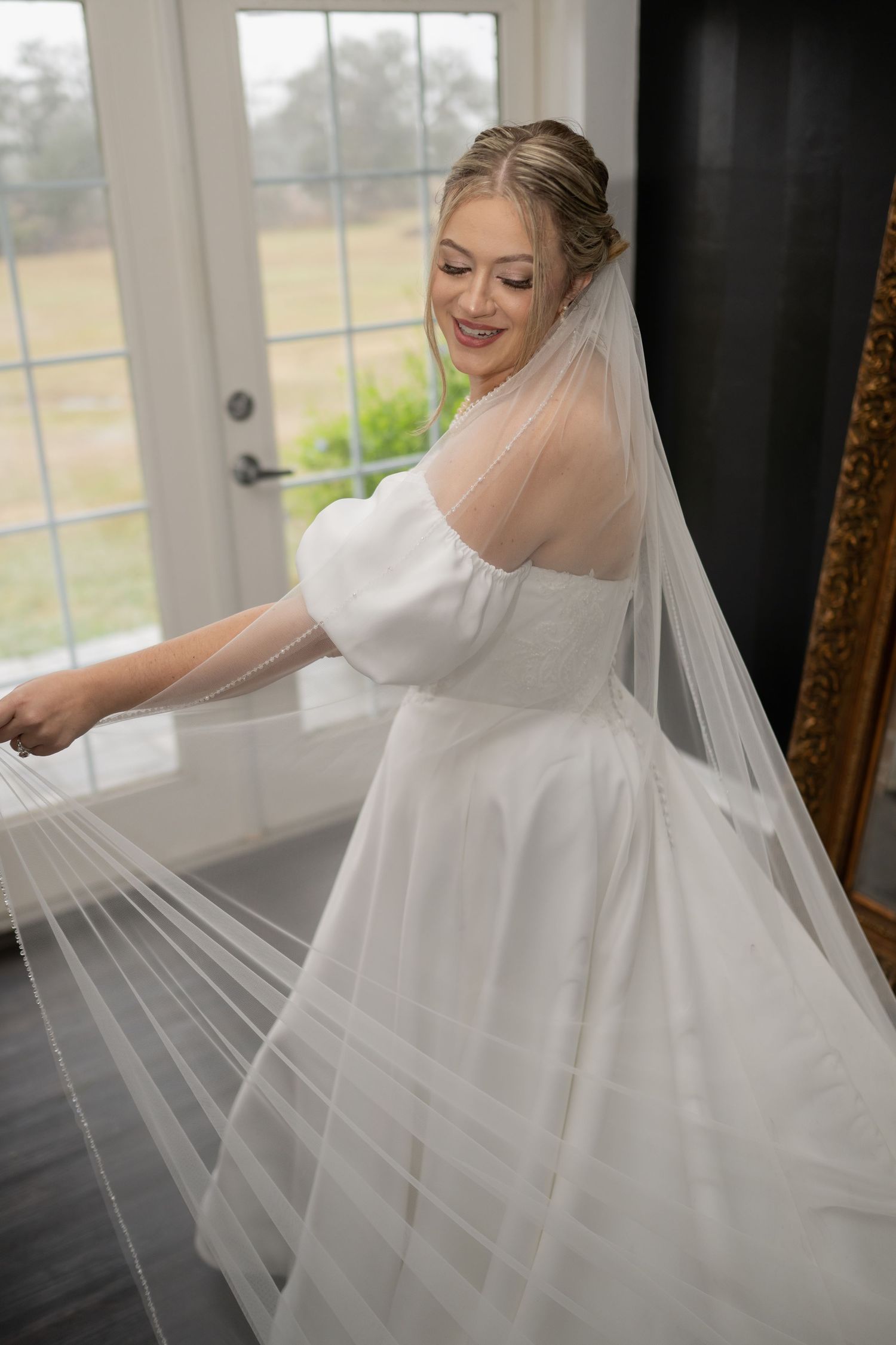 Smiling bride in white gown with puff sleeves and long veil posing near French doors on her wedding day.