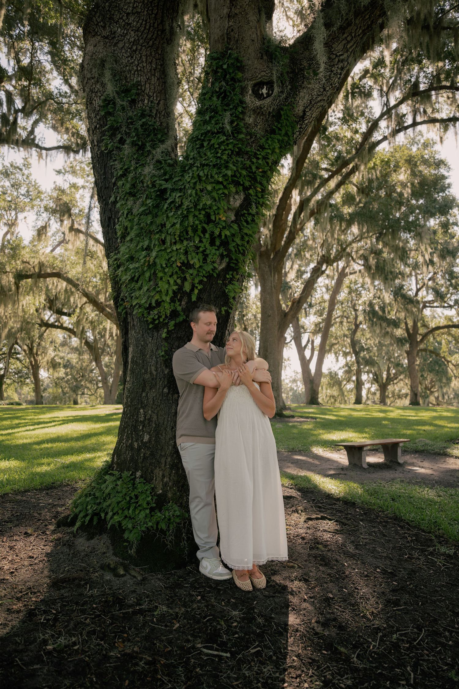 Couple embracing under a large ivy-covered oak tree in a lush green park, man in tan shirt hugging woman in white dress.