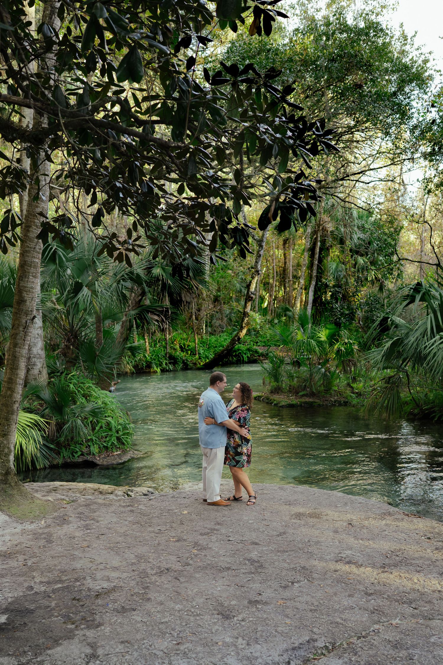 Couple embracing romantically by a lush green river surrounded by tropical trees and palms in a serene outdoor setting.