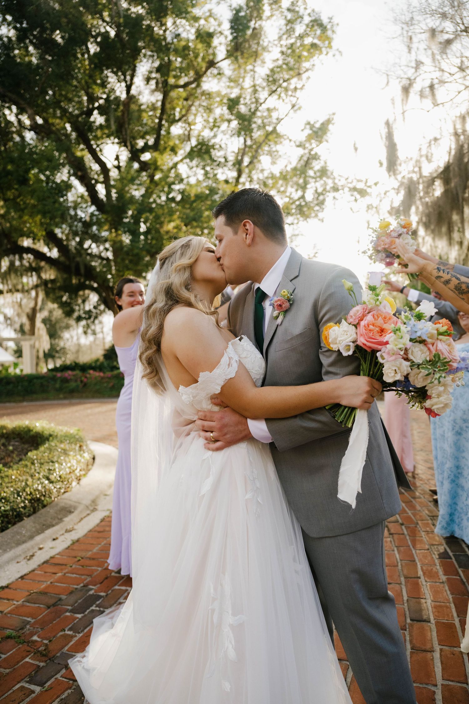 Bride and groom share a first kiss on brick pathway surrounded by lush trees and colorful wedding party.
