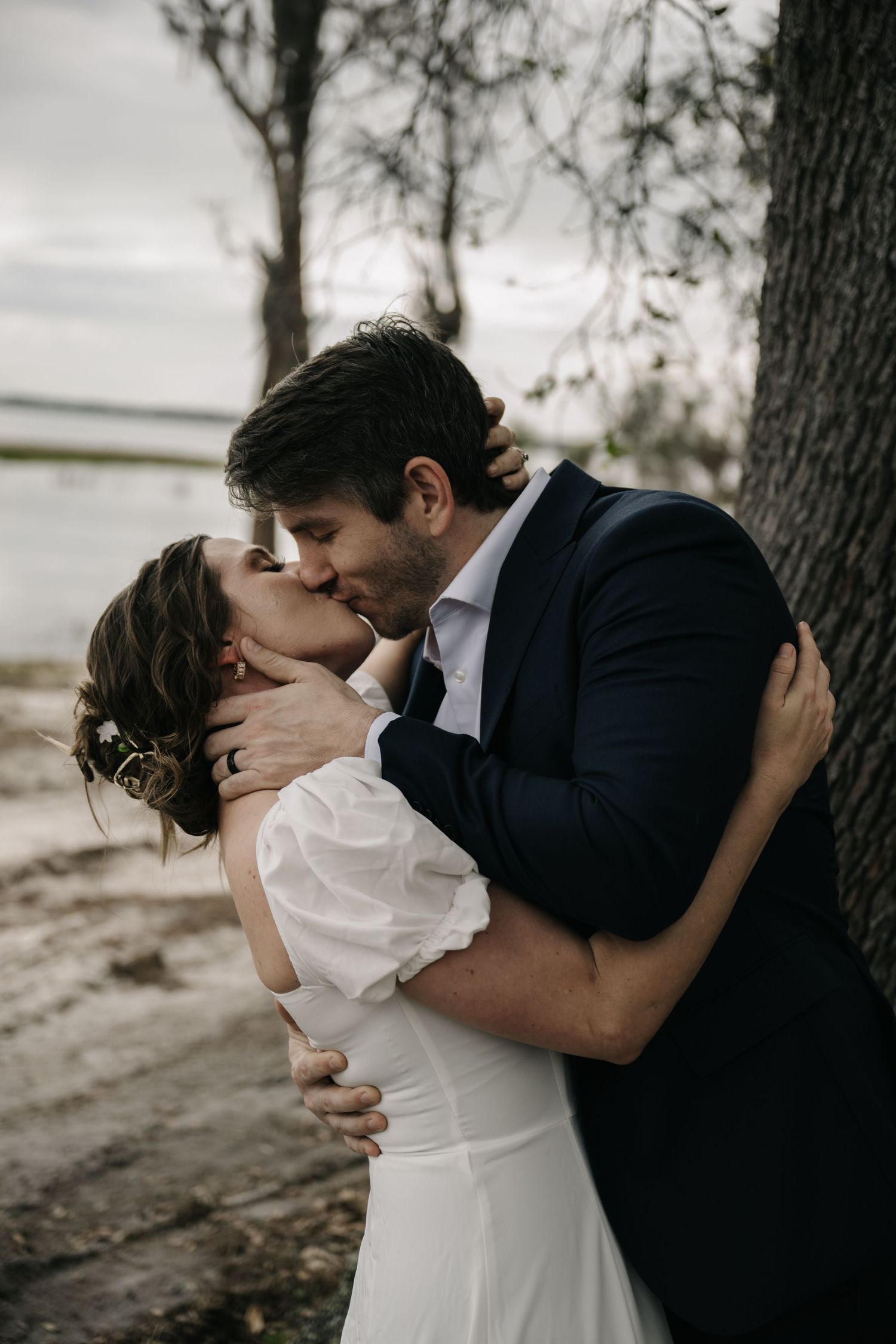 Romantic wedding couple shares intimate kisses against trees in a natural outdoor setting.