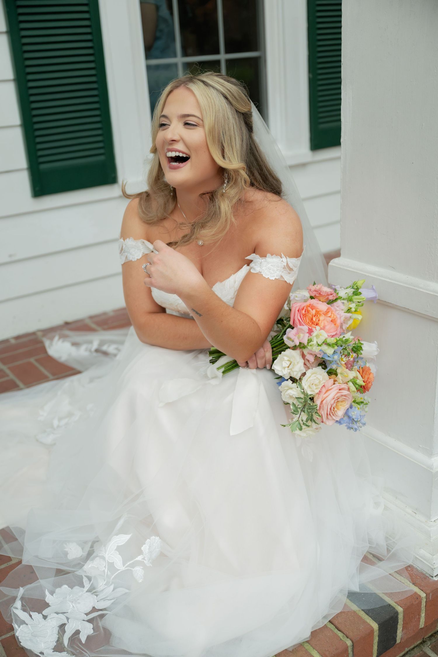 Bride in white tulle dress laughs joyfully while sitting on brick steps with floral bouquet.